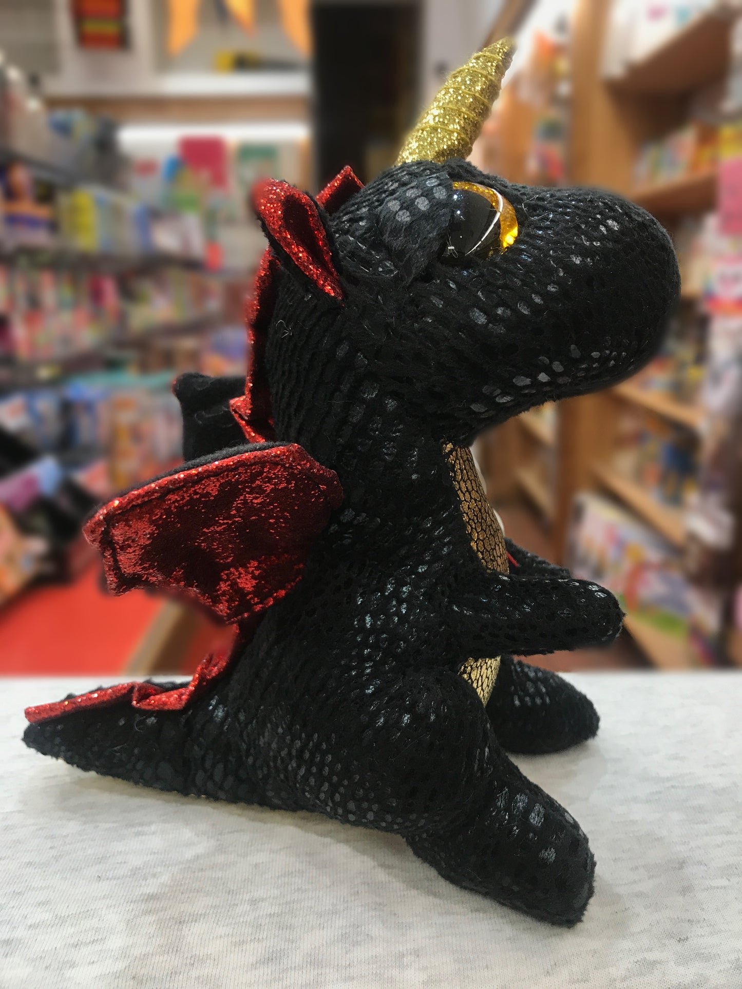 Ty Beanie Boo Grindal dragon plush with black scales, red sparkly wings, gold horn, and golden eyes in a toy store setting