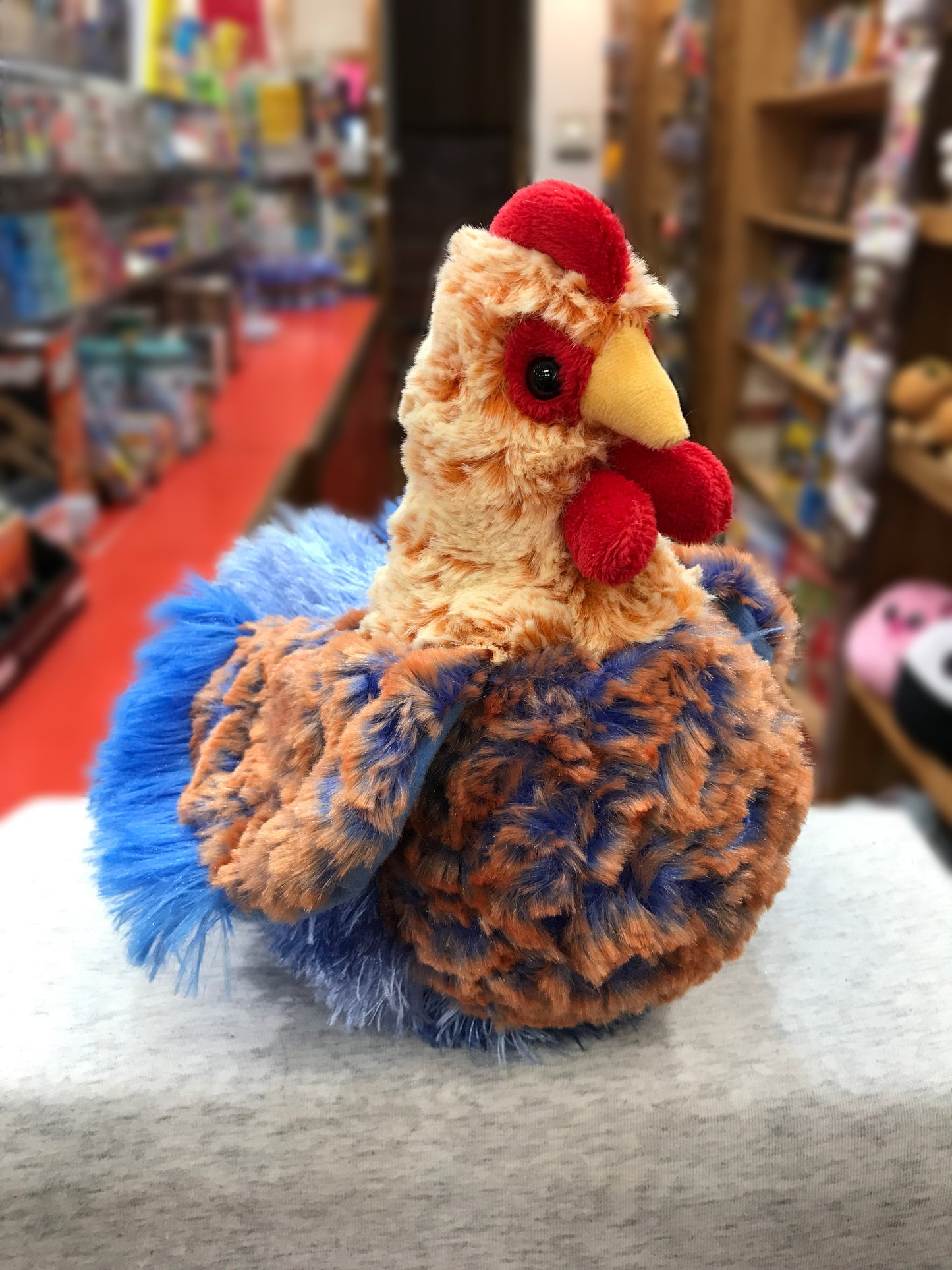 Colorful plush Blue Lace Hen stuffed animal with vibrant red comb and wattle, displayed in toy store setting