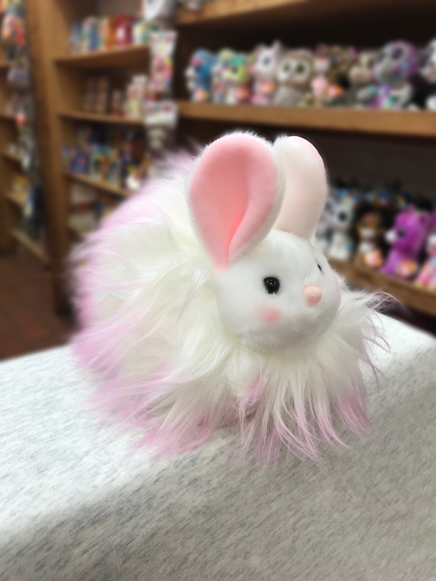 Cute pink and white Douglas Puff Bunny plush toy with fluffy fur, displayed in a toy store with other stuffed animals on shelves