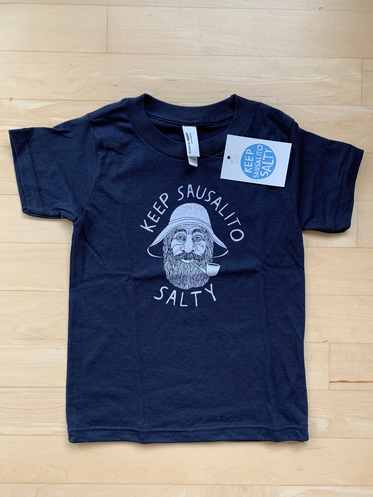 Navy kids' t-shirt with "Keep Sausalito Salty" slogan and sailor illustration, tag attached, on wooden floor