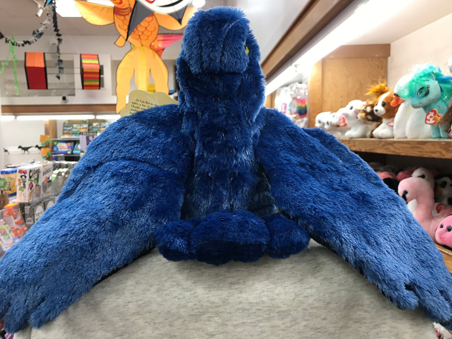 Vibrant blue macaw puppet with movable wings in toy store display, surrounded by colorful plush animals and decorations