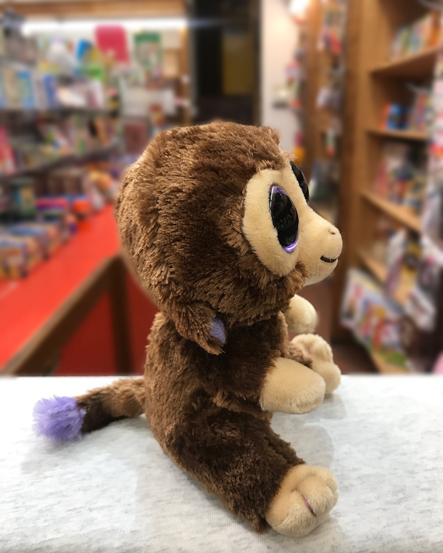 Ty Beanie Boo Coconut Monkey plush toy with large purple eyes, sitting on display in a toy store surrounded by colorful shelves