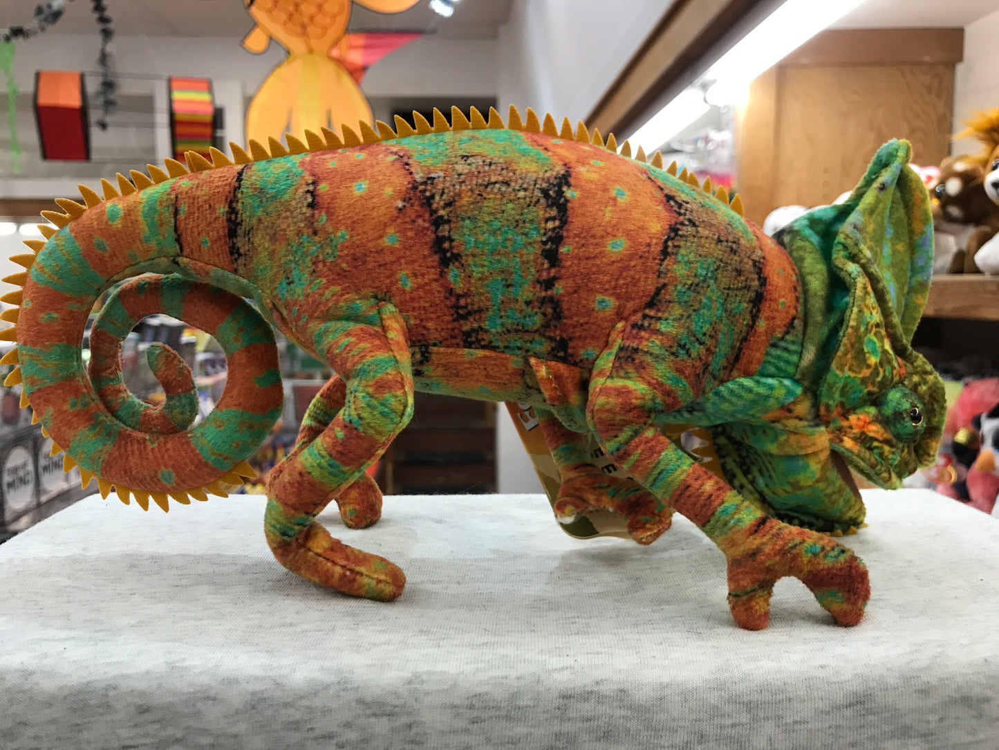 Colorful Folkmanis chameleon puppet with detailed fabric design, movable mouth, and curled tail on display in toy store