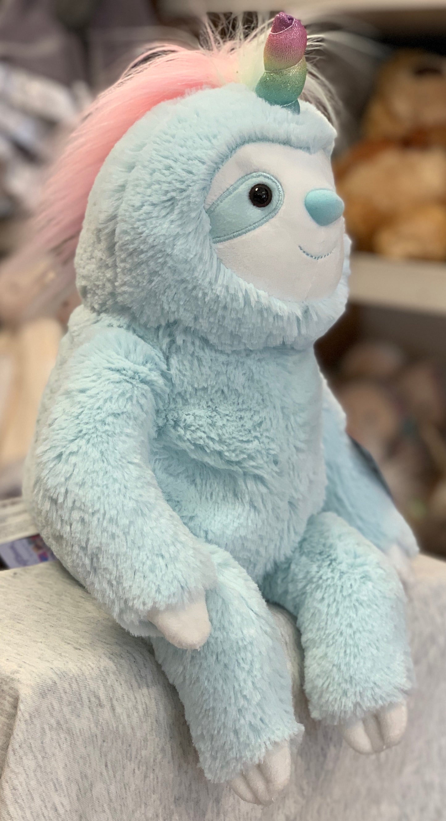 Gund Slothicorn plush toy with light blue fur, pink mane, and iridescent rainbow horn sitting on fabric surface