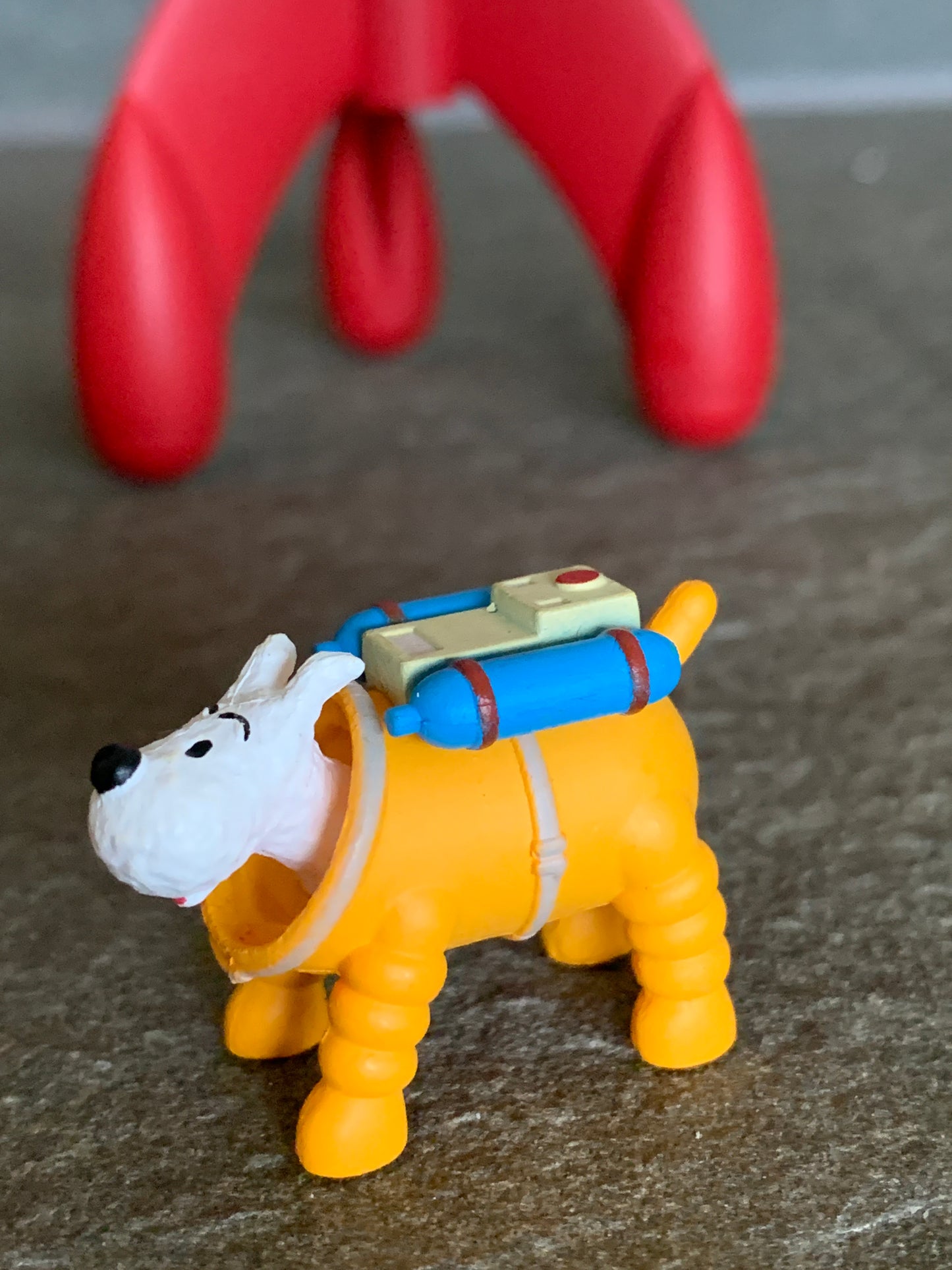 Snowy cosmonaut mini figure in a yellow spacesuit with a blue oxygen tank on his back, standing in front of a blurred red moon rocket.