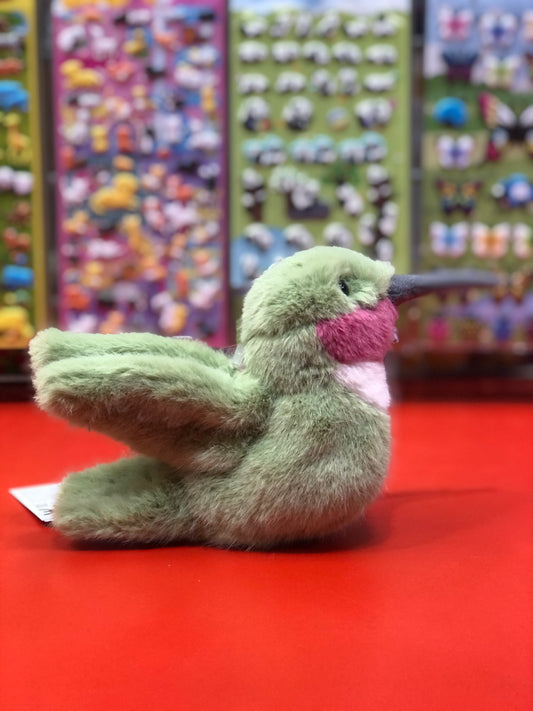 Plush green hummingbird toy with pink throat on red surface, colorful plush animal display in background