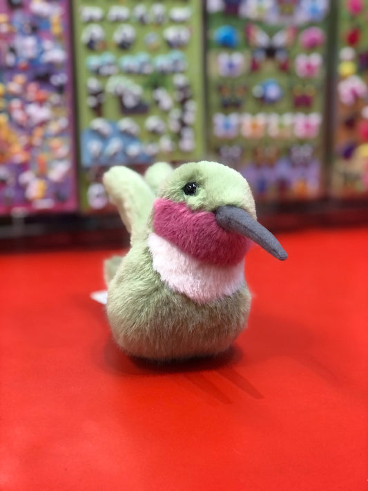 Plush hummingbird toy with green body, pink throat, and long beak on red surface, colorful toy display in background