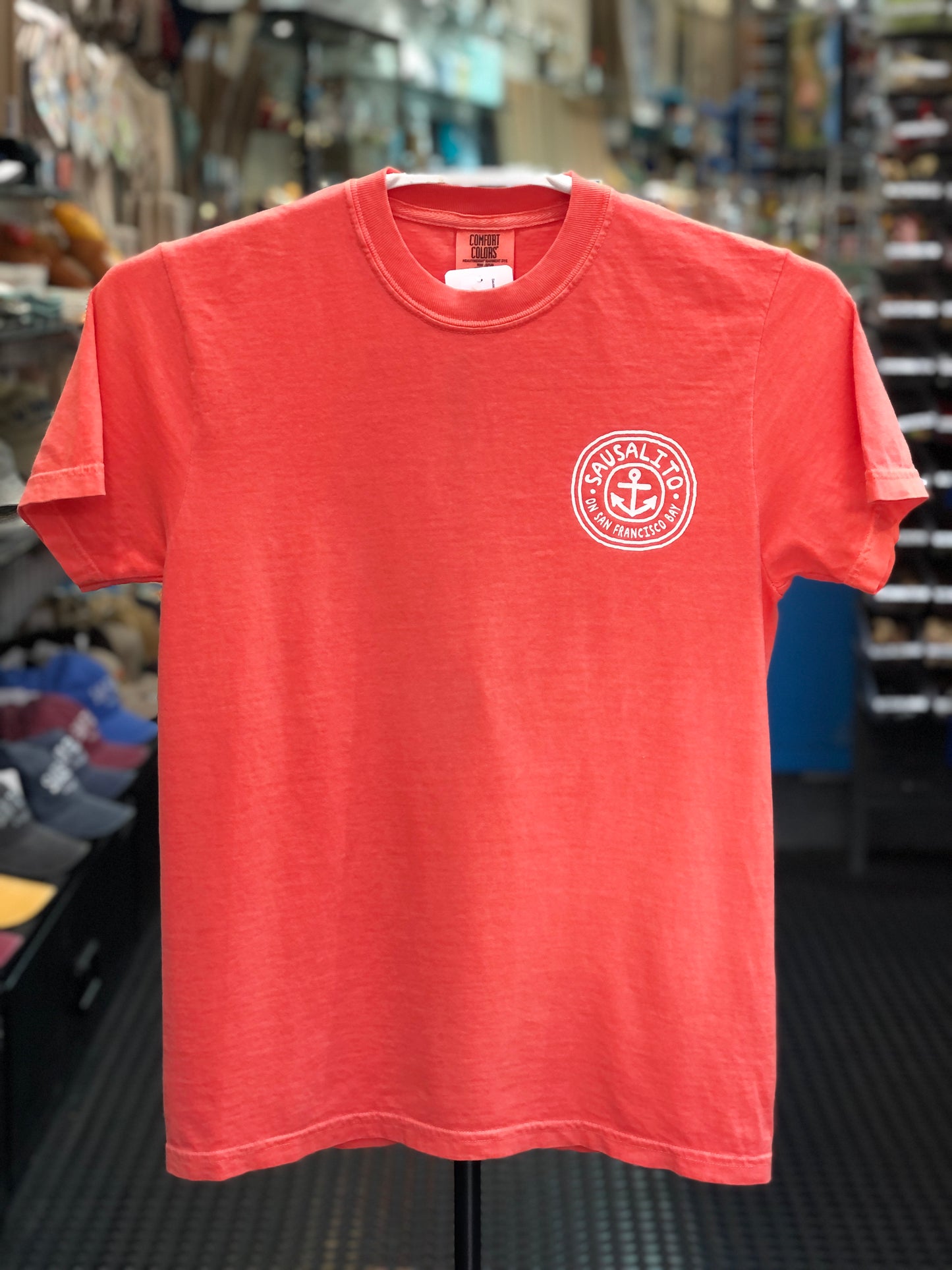 Orange Comfort Colors t-shirt with a circular logo featuring an anchor and text on a hanger in a store. 