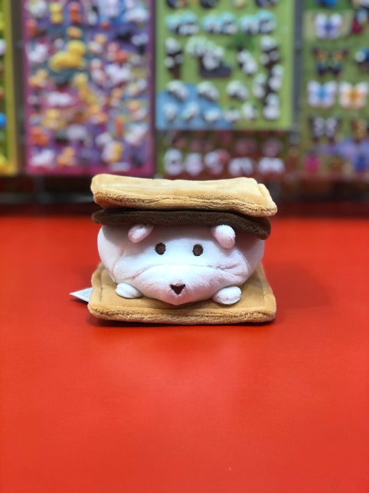 Cute S'more Bear plush toy with white marshmallow bear between graham crackers, displayed on red surface with colorful background