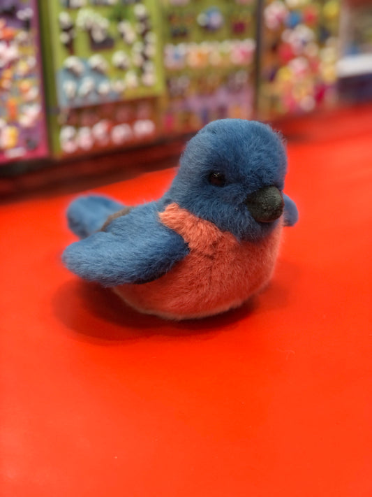 Adorable Douglas Bertie blue bird plush toy with orange belly sitting on red surface, colorful toy display in background