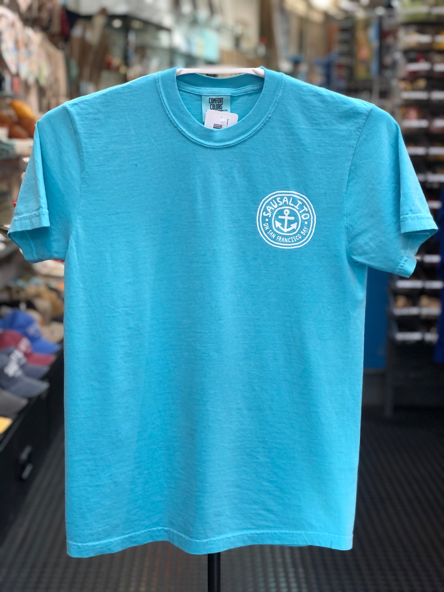 Light blue Comfort Colors t-shirt with a circular logo featuring an anchor and text on a hanger in a store. 
