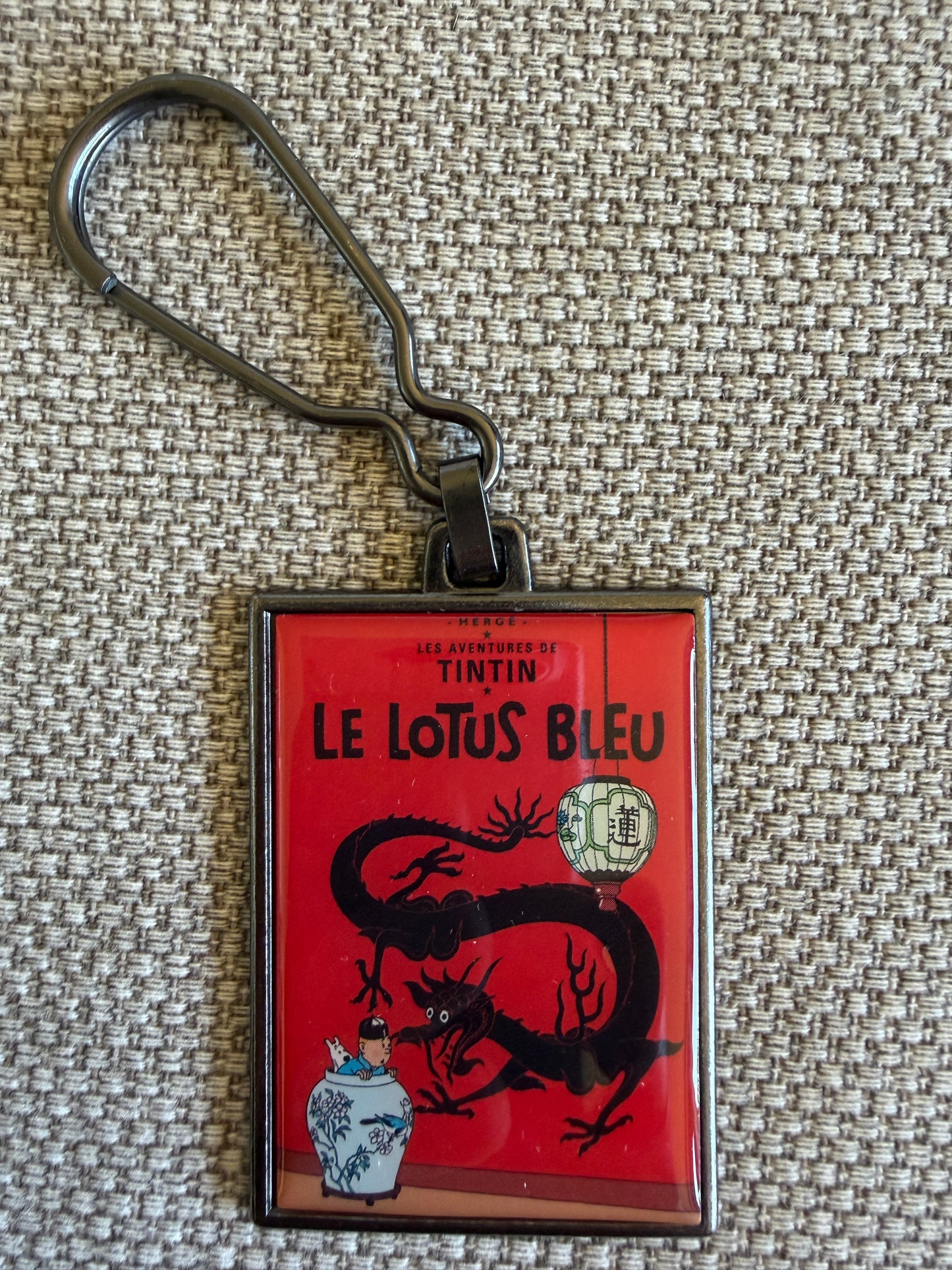 A front close-up view of an officially authorized, rectangular metal Tintin keyring resting on a textured beige fabric surface. The keyring features the vibrant enamel front cover art from "The Blue Lotus," showing a startled Tintin peaking out of a blue and white porcelain vase, with a prominent, ferocious black dragon stretching across a striking red background.