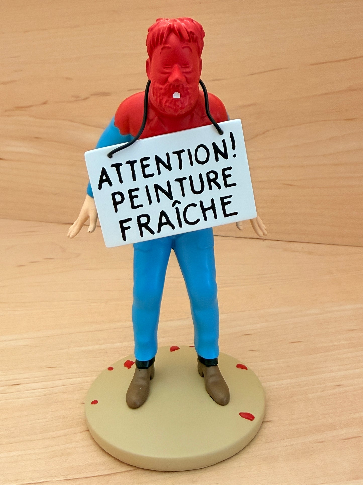 12 cm hand-painted resin figure of a distressed Captain Haddock from Hergé's Tintin comic Destination Moon. His face, shoulders, and hair are entirely covered in bright red wet paint, and a white placard hanging around his neck reads "ATTENTION! PEINTURE FRAÎCHE" in black capital letters.
