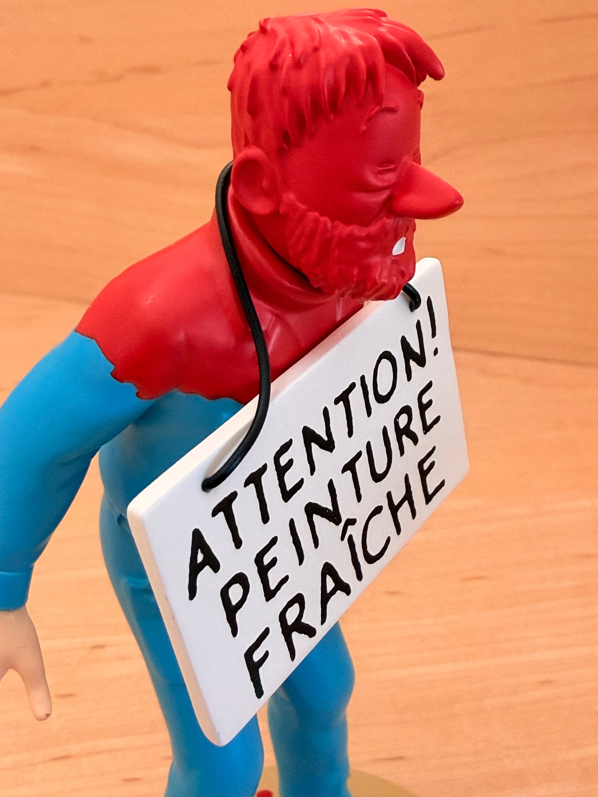 A profile view of a 12 cm hand-painted resin figure showing Captain Haddock covered in bright red wet paint across his head, face, and shoulders. A white placard hanging by a black string around his neck reads "ATTENTION! PEINTURE FRAÎCHE" in bold black lettering, resting against his classic blue sweater.