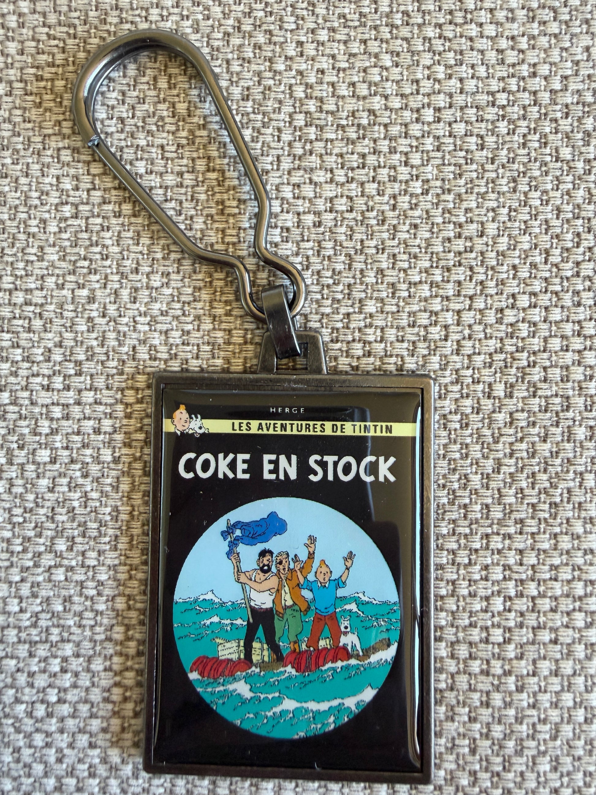A rectangular keychain displaying the French book cover art for Hergé's Tintin adventure "Coke en Stock." The illustration shows Tintin, Captain Haddock waving his shirt on a stick, and pilot Skut waving on a wooden raft in the middle of a vibrant blue ocean.