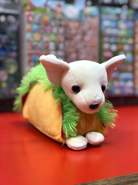 Adorable plush Chihuahua in taco costume with fluffy green lettuce fringe, sitting on red surface in toy store display