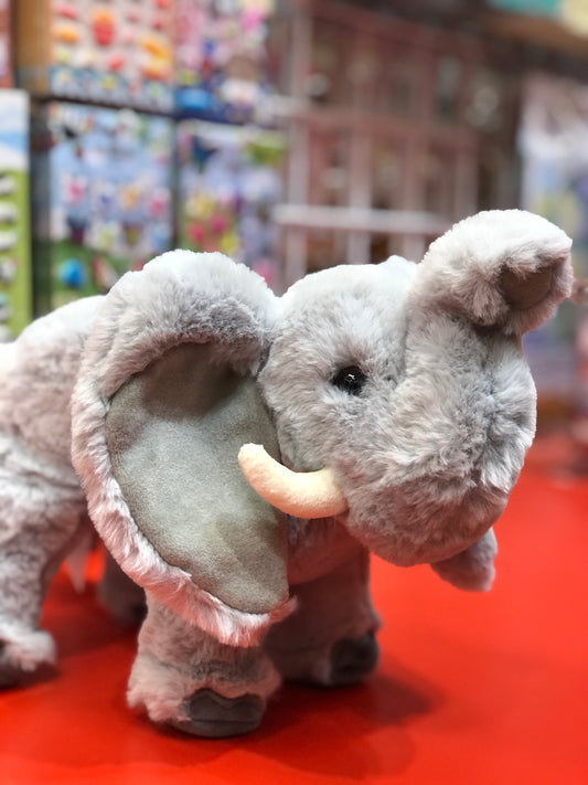 Soft gray elephant plush toy with floppy ears and upraised trunk on red display, colorful toy shelves in background