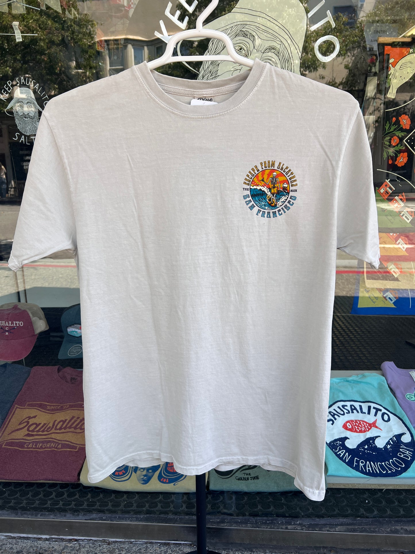 Front view of a cement gray garment-dyed t-shirt on a hanger against a store window background, featuring a small, vibrant circular chest graphic on the left pocket showing a surfing skeleton and the text "ESCAPE FROM ALCATRAZ SAN FRANCISCO".