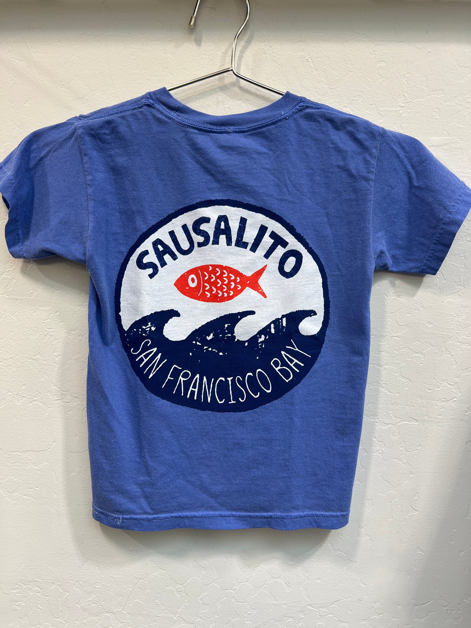 Blue kids t-shirt with Sausalito logo featuring red fish and waves, San Francisco Bay text, hanging on hanger