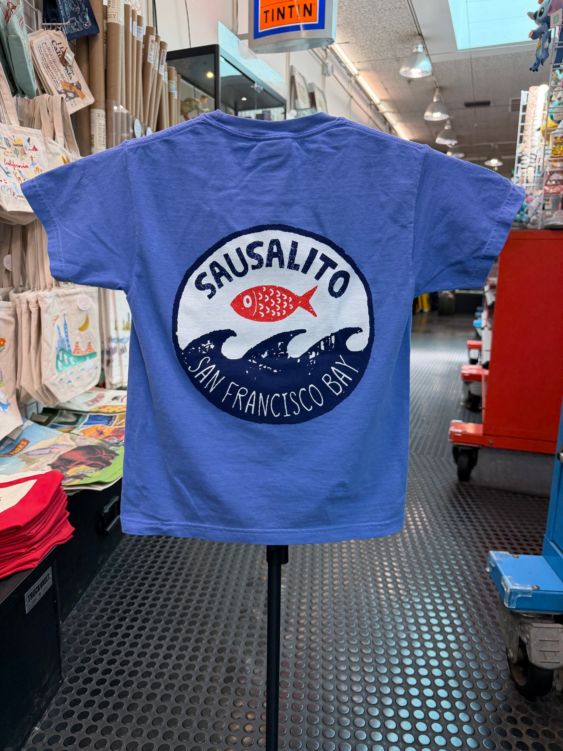 The back of a flo blue short-sleeve kids T-shirt displayed on a stand inside a retail store. The shirt features a large circular graphic on the back with "Sausalito" written in a bold arch over a bright red fish, which sits above stylized navy blue ocean waves. Underneath the waves, the text reads "San Francisco Bay."