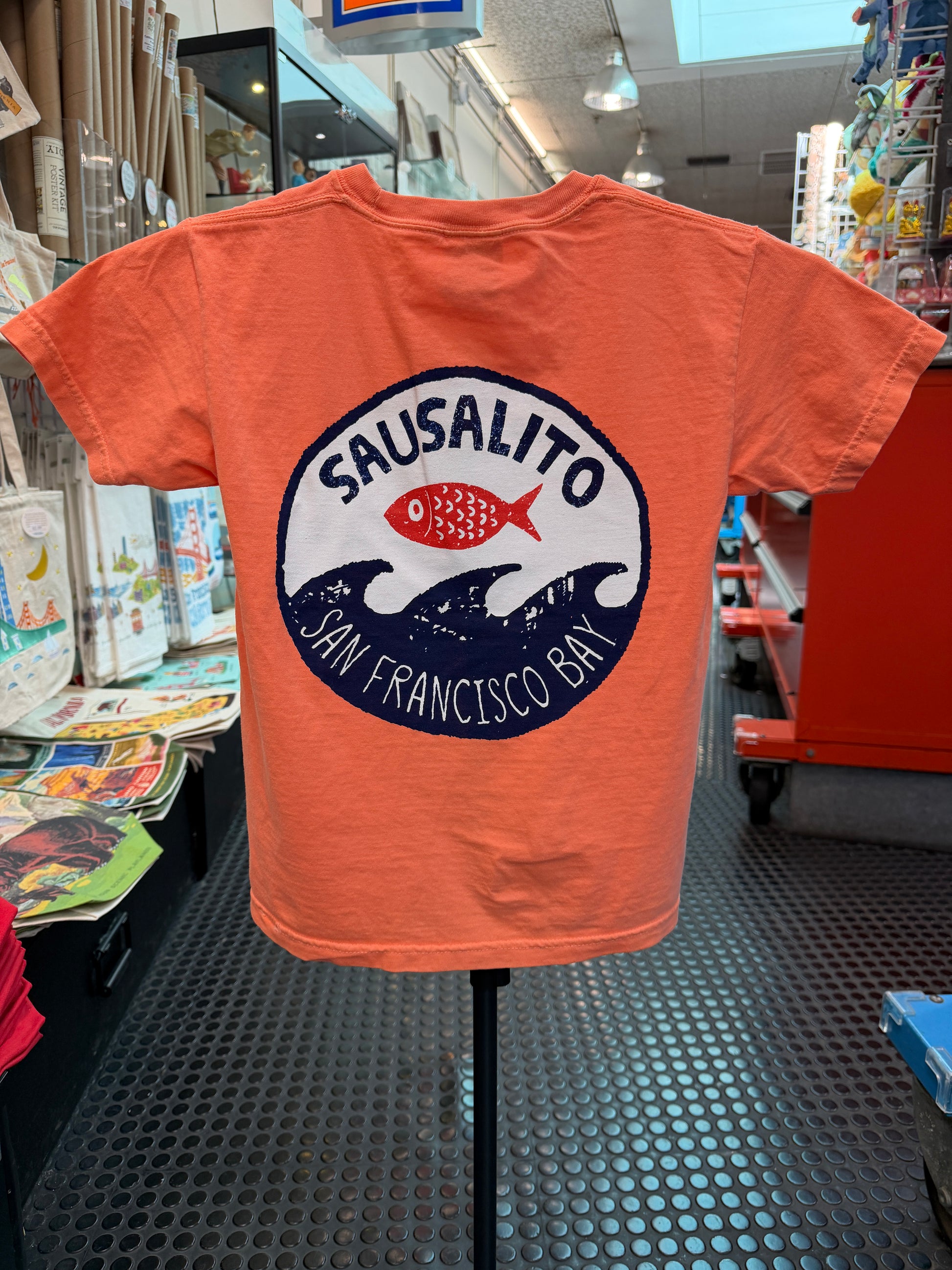 he back of a melon orange short-sleeve kids T-shirt displayed on a stand inside the Sausalito Ferry Co. retail store. The shirt features a large circular graphic on the back with "Sausalito" written in a bold arch over a bright red fish, which sits above stylized navy blue ocean waves. Underneath the waves, the text reads "San Francisco Bay."