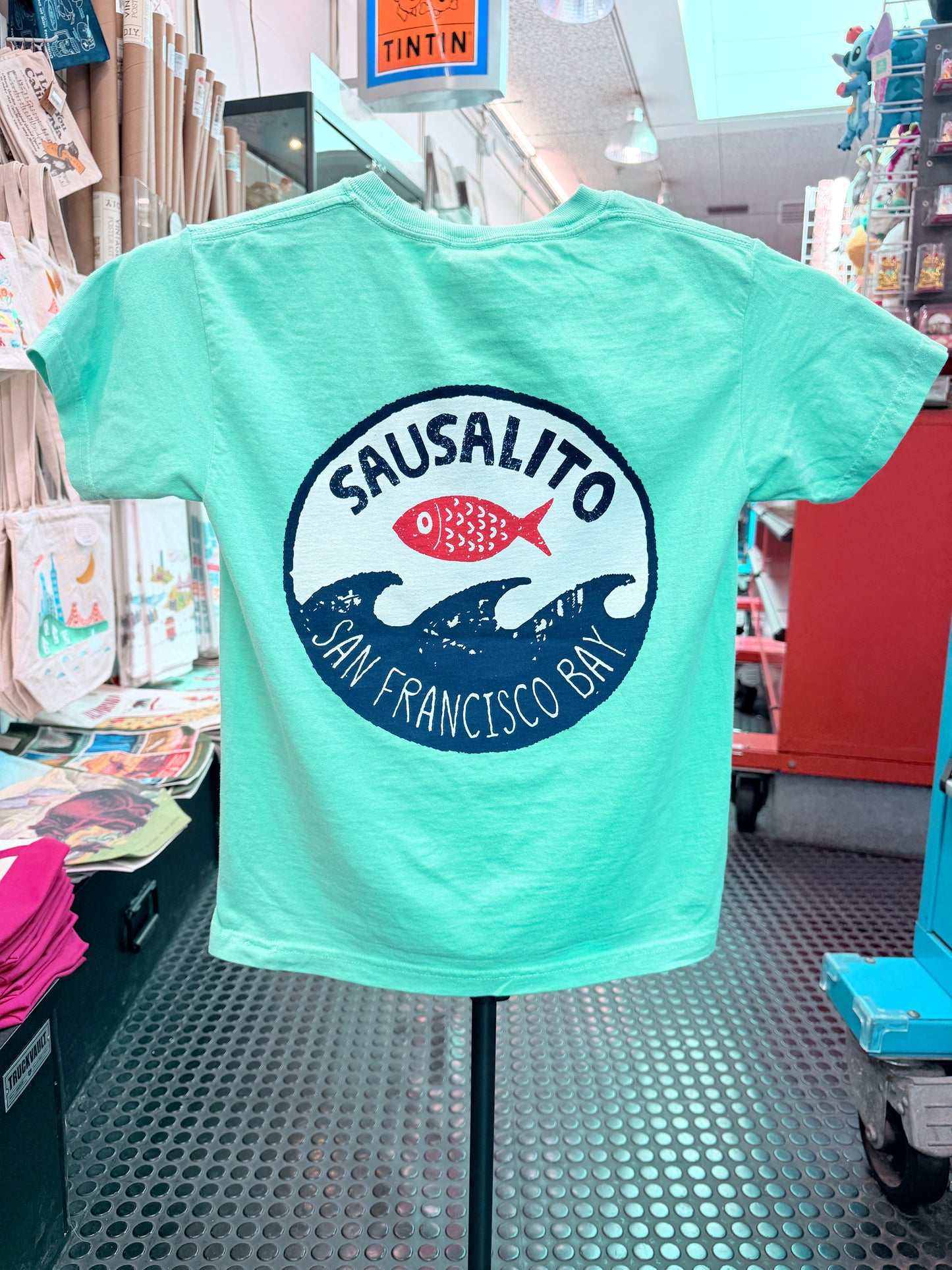 The back of a mint green short-sleeve kids T-shirt displayed on a stand inside the Sausalito Ferry Co. retail store. The shirt features a large circular graphic on the back with "Sausalito" written in a bold arch over a bright red fish, which sits above stylized navy blue ocean waves. Underneath the waves, the text reads "San Francisco Bay."