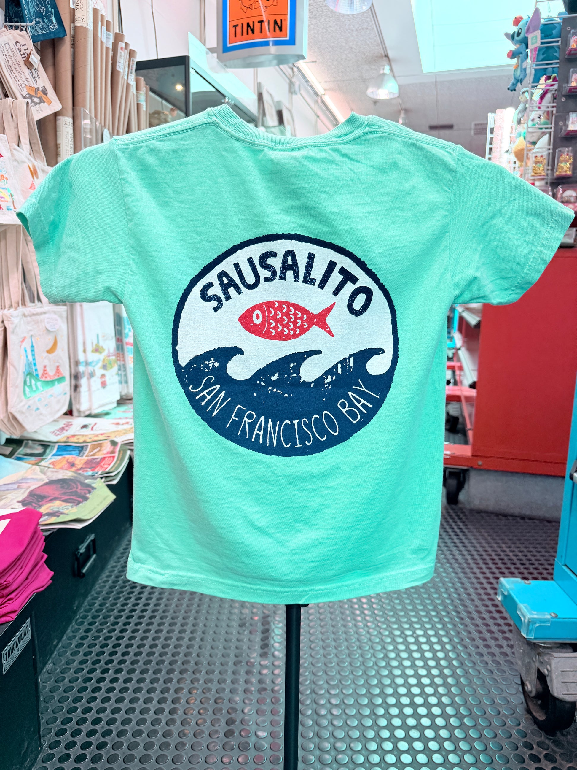 The back of a mint green short-sleeve kids T-shirt displayed on a stand inside the Sausalito Ferry Co. retail store. The shirt features a large circular graphic on the back with "Sausalito" written in a bold arch over a bright red fish, which sits above stylized navy blue ocean waves. Underneath the waves, the text reads "San Francisco Bay."