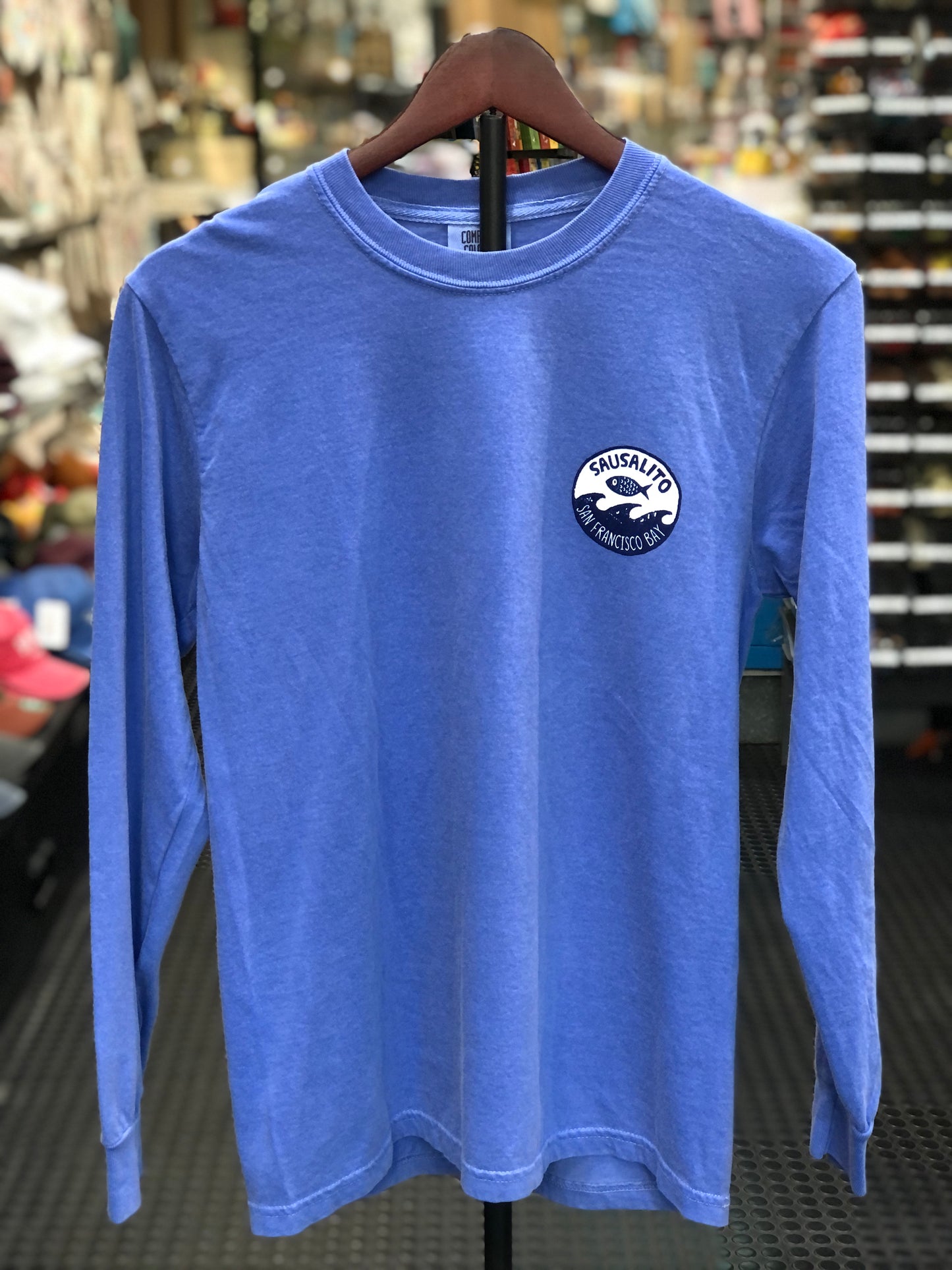 Front view of a flo blue garment-dyed long-sleeve t-shirt on a hanger in a store setting, featuring a small circular chest graphic on the left side with a stylized fish leaping over waves and the text "SAUSALITO SAN FRANCISCO BAY".