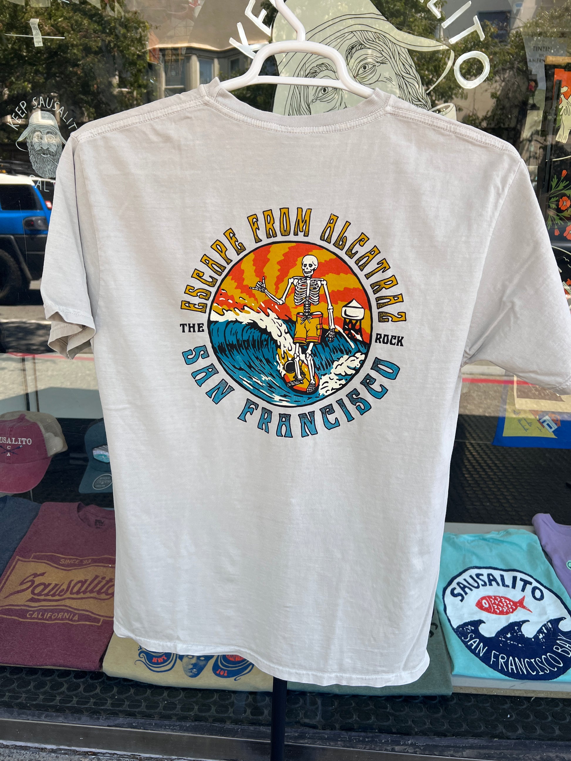 Back view of a cement gray garment-dyed t-shirt hanging on a rack, featuring a large circular graphic of a surfing skeleton with a ball and chain on a wave, set against a vibrant orange and yellow sunset with the text "ESCAPE FROM ALCATRAZ - THE ROCK - SAN FRANCISCO".