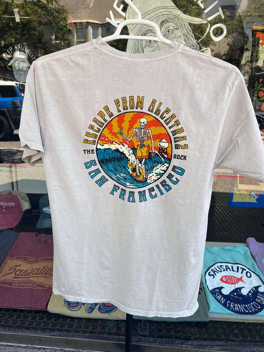 Back view of a cement gray garment-dyed t-shirt hanging on a rack, featuring a large circular graphic of a surfing skeleton with a ball and chain on a wave, set against a vibrant orange and yellow sunset with the text "ESCAPE FROM ALCATRAZ - THE ROCK - SAN FRANCISCO".