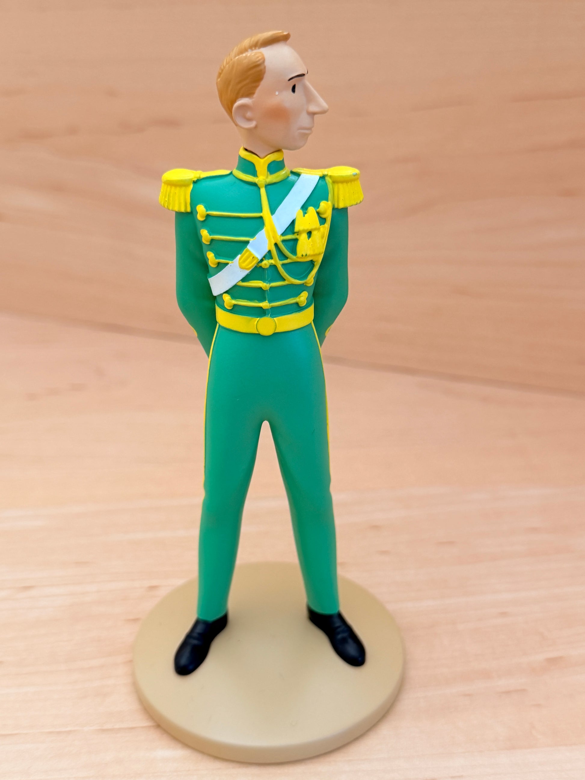 A 12 cm hand-painted resin figure representing the creator Hergé in a cameo appearance from the Tintin album King Ottokar’s Sceptre. The figure is depicted standing at attention in a bright green Syldavian full dress uniform with a yellow collar, gold epaulets, gold braided detailing across the chest, a light blue ceremonial sash, and black shoes.