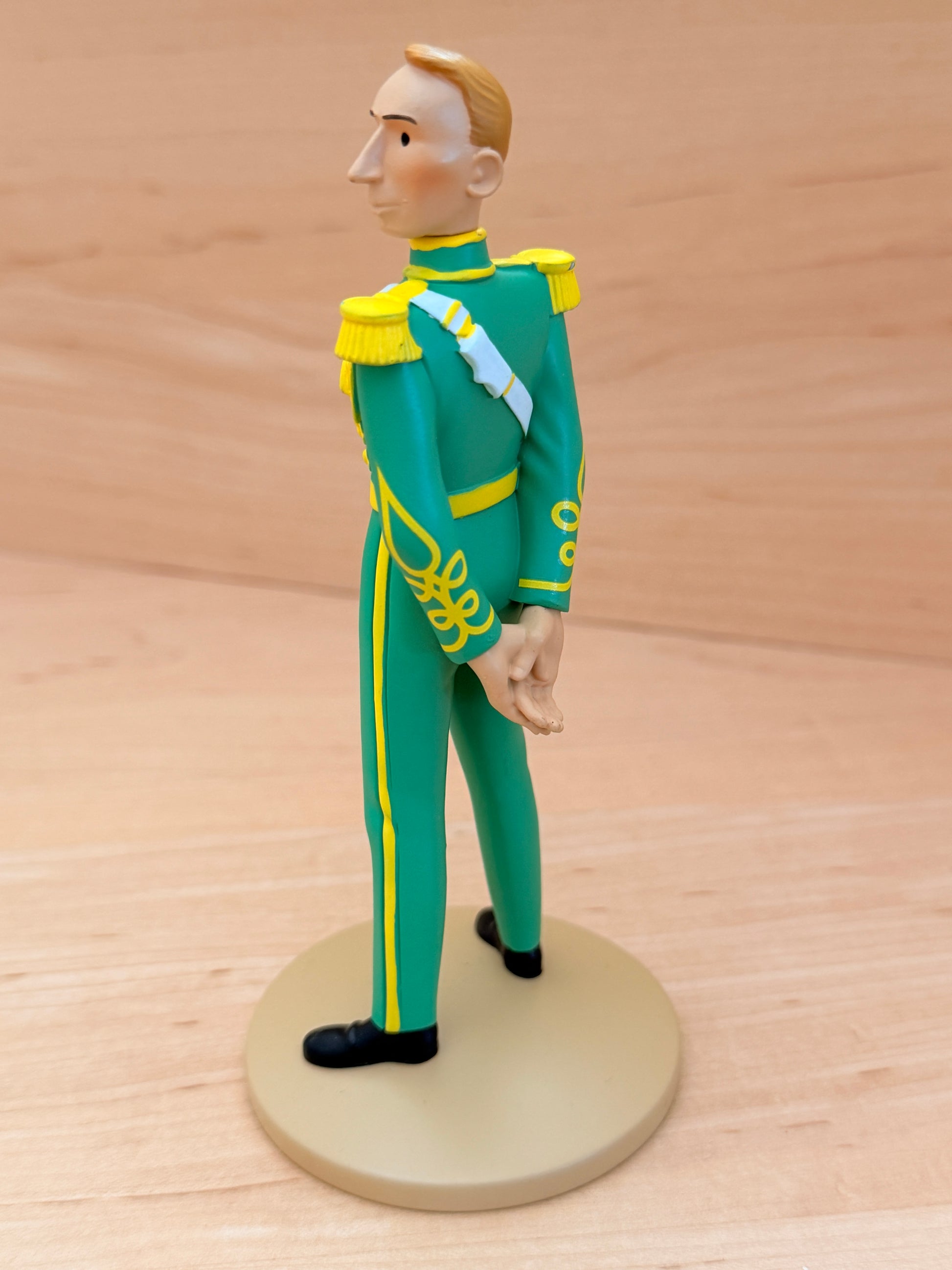 A side and rear profile view of a 12 cm hand-painted resin figure representing the creator Hergé in a Syldavian full dress uniform from King Ottokar’s Sceptre. The figure stands with hands clasped behind his back, showcasing the bright green uniform with yellow trousers stripes, gold fringed shoulder epaulets, and a light blue ceremonial sash crossing down the back.