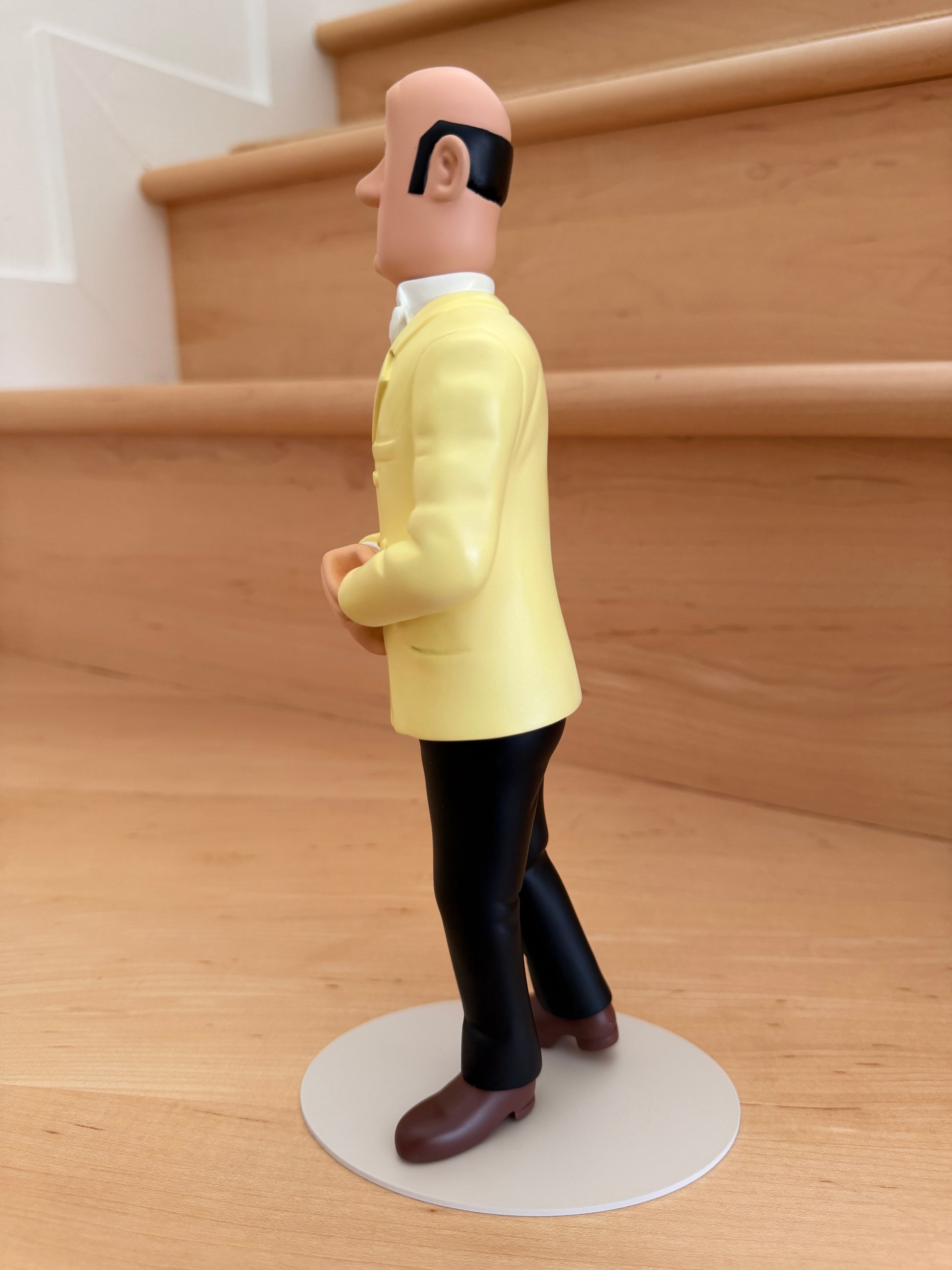 Left side profile view of the Tintinimaginatio Icons collection resin figurine of Nestor, the butler of Marlinspike Hall. He is depicted from the side with a calm expression, wearing his classic yellow double-breasted suit jacket, black trousers, and a white bowtie, standing on a thin white circular base against a background of wooden steps.