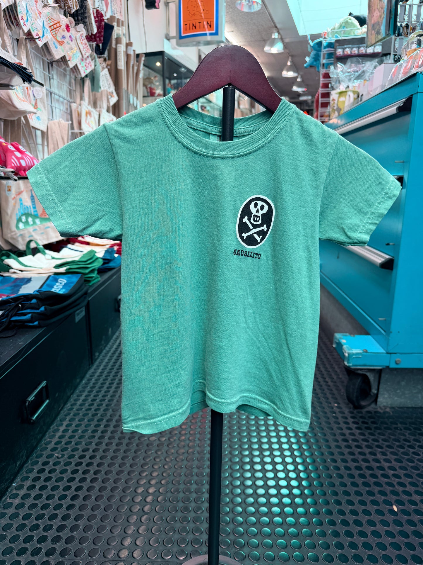 A mint green or seafoam short-sleeve kid's t-shirt hanging on a display hanger. It features a black circular chest graphic with a white skull and crossbones and the text 'SAUSALITO' printed underneath the bones.