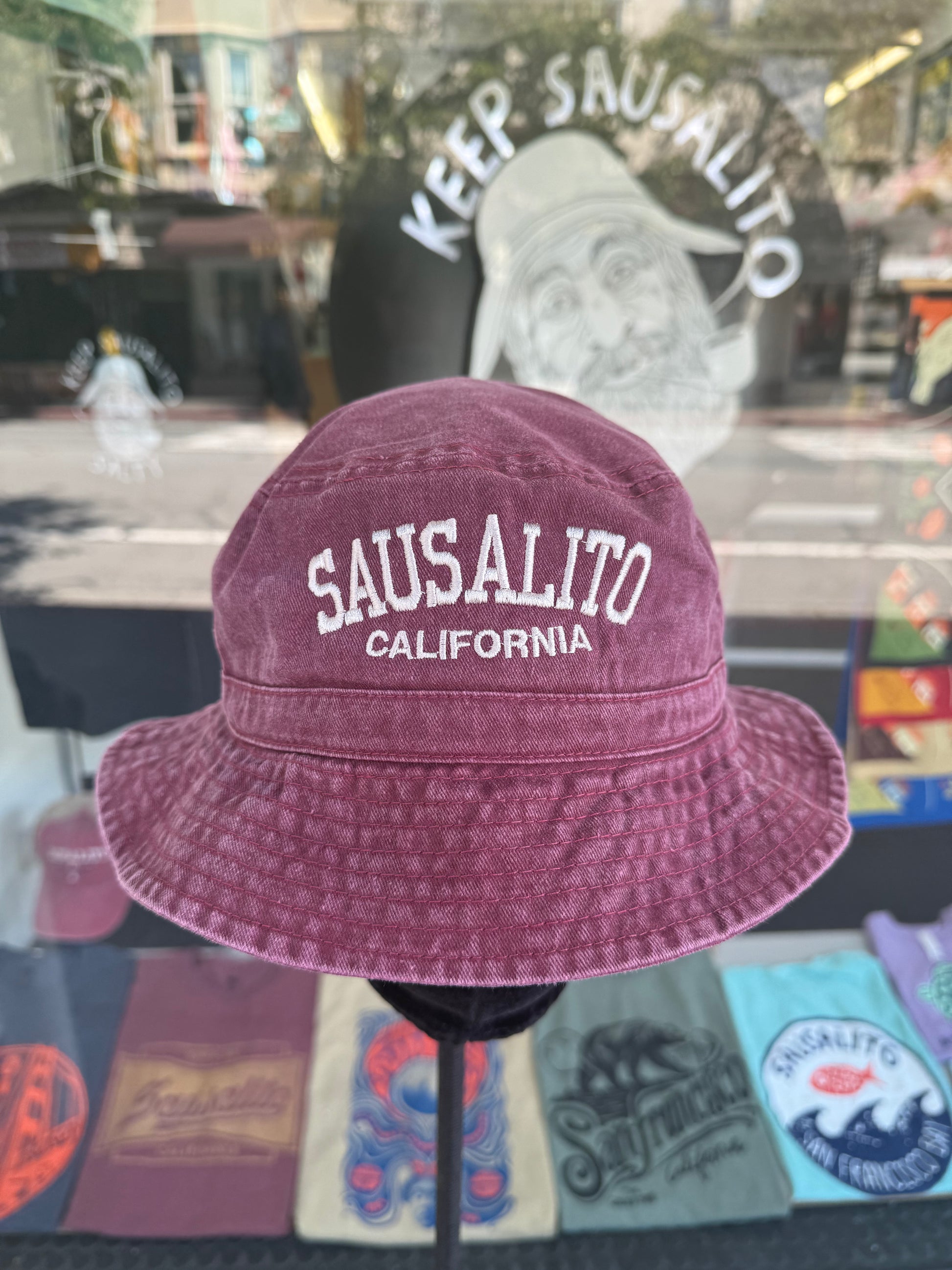A front view of a burgundy, over-washed 100% cotton bucket hat displayed on a black mannequin stand in front of a store window. The front features the words "SAUSALITO" cleanly embroidered in white block letters arched over "CALIFORNIA" in smaller white text. The window reflection shows a vintage style illustration under the arched words "KEEP SAUSALITO SALTY".