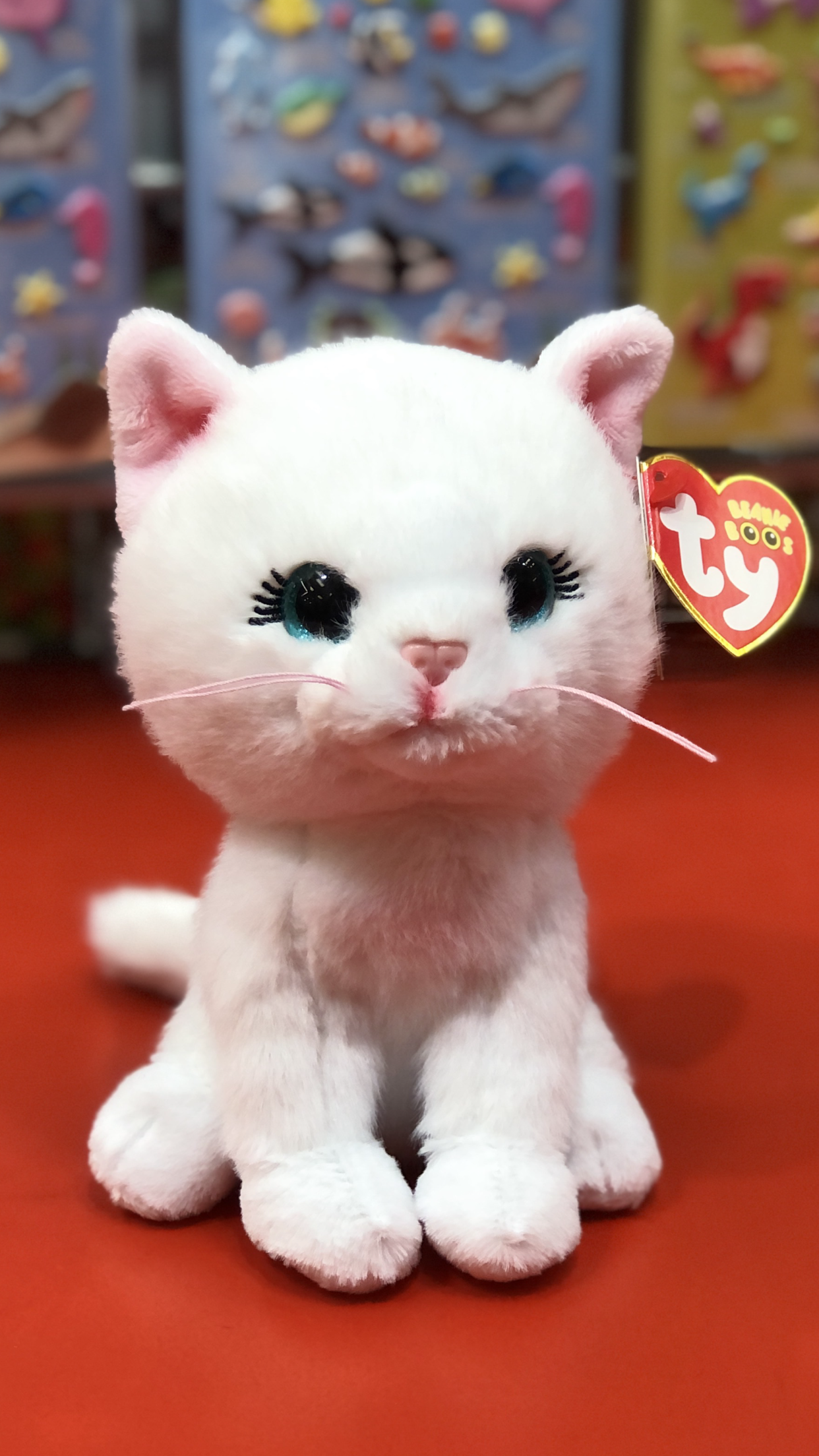 White Ty plush cat toy with black eyes and pink nose on a red surface, with a colorful background.