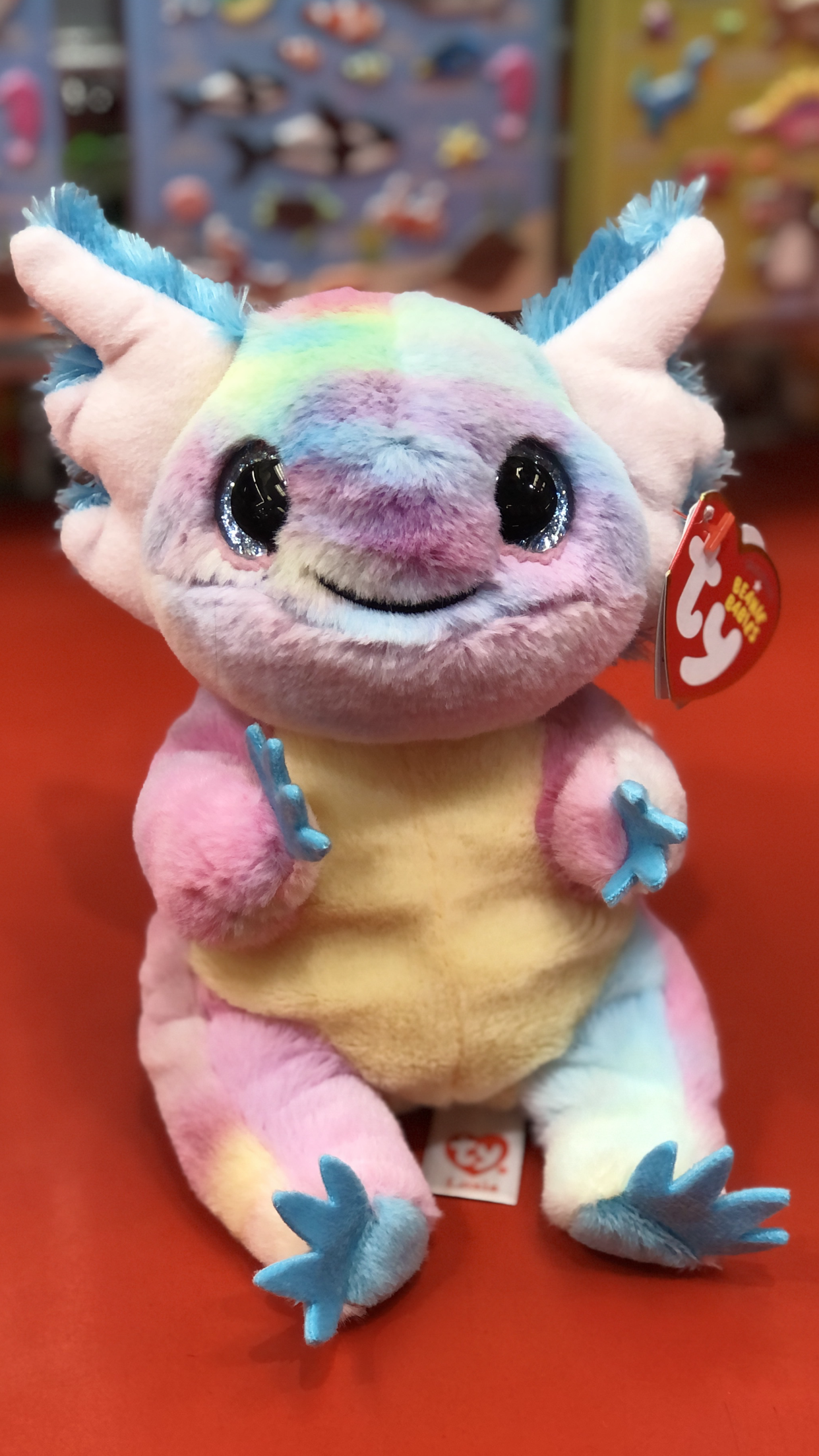 Ty beanie babies style rainbow axolotl plush with a smiling face on a red surface.