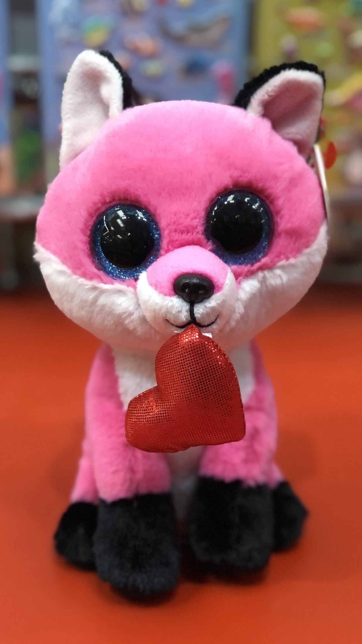 Ty pink fox plush with black ears and black feet with a red heart sitting on a red surface. 