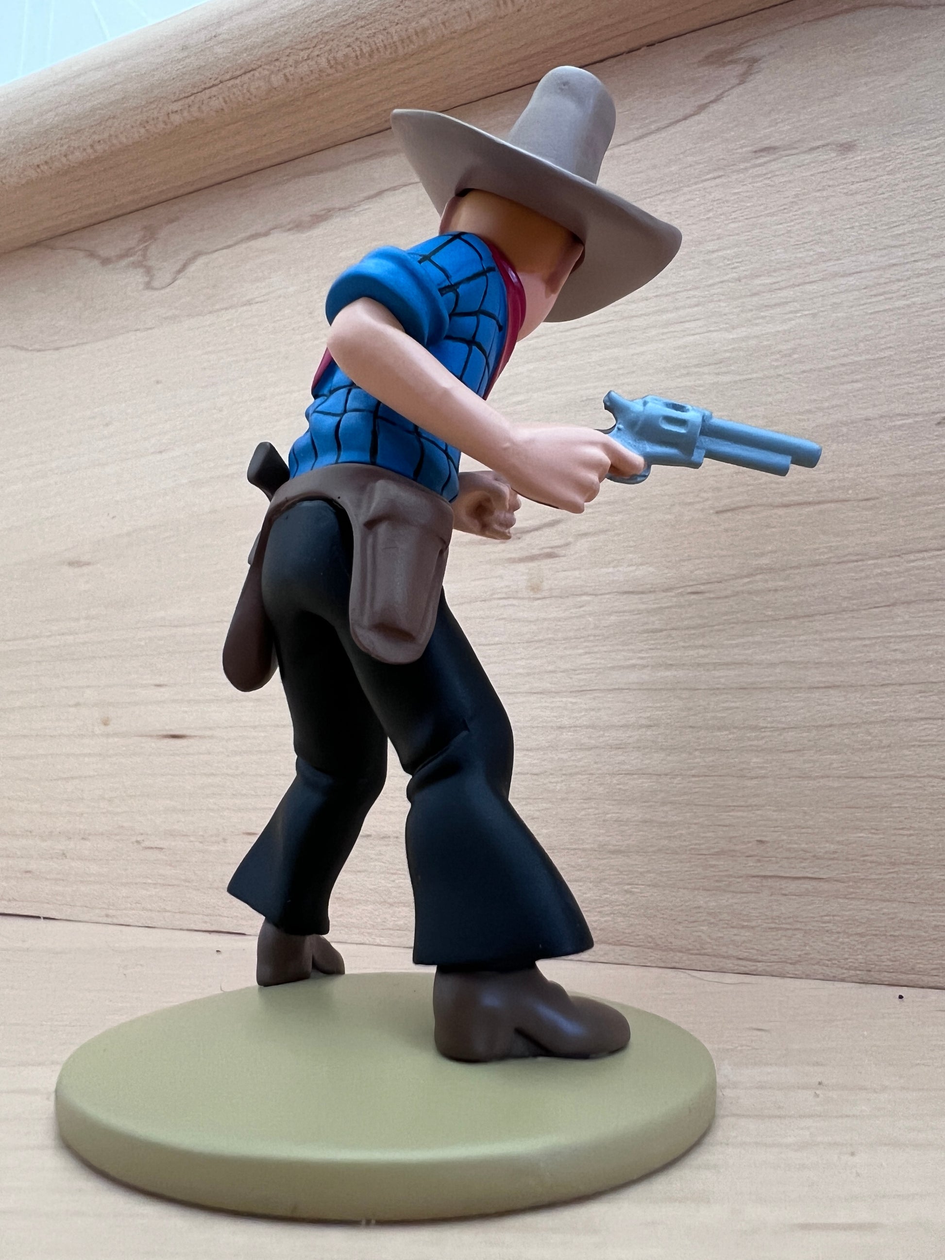 Side-profile view of the Tintin in America polyresin mini figure, showing Tintin from the right side in a blue-and-white checkered shirt, black pants with a brown holster and gun belt, and a wide-brimmed grey cowboy hat, captured in a dynamic crouching stance while holding a silver revolver pistol on a round olive-green base.