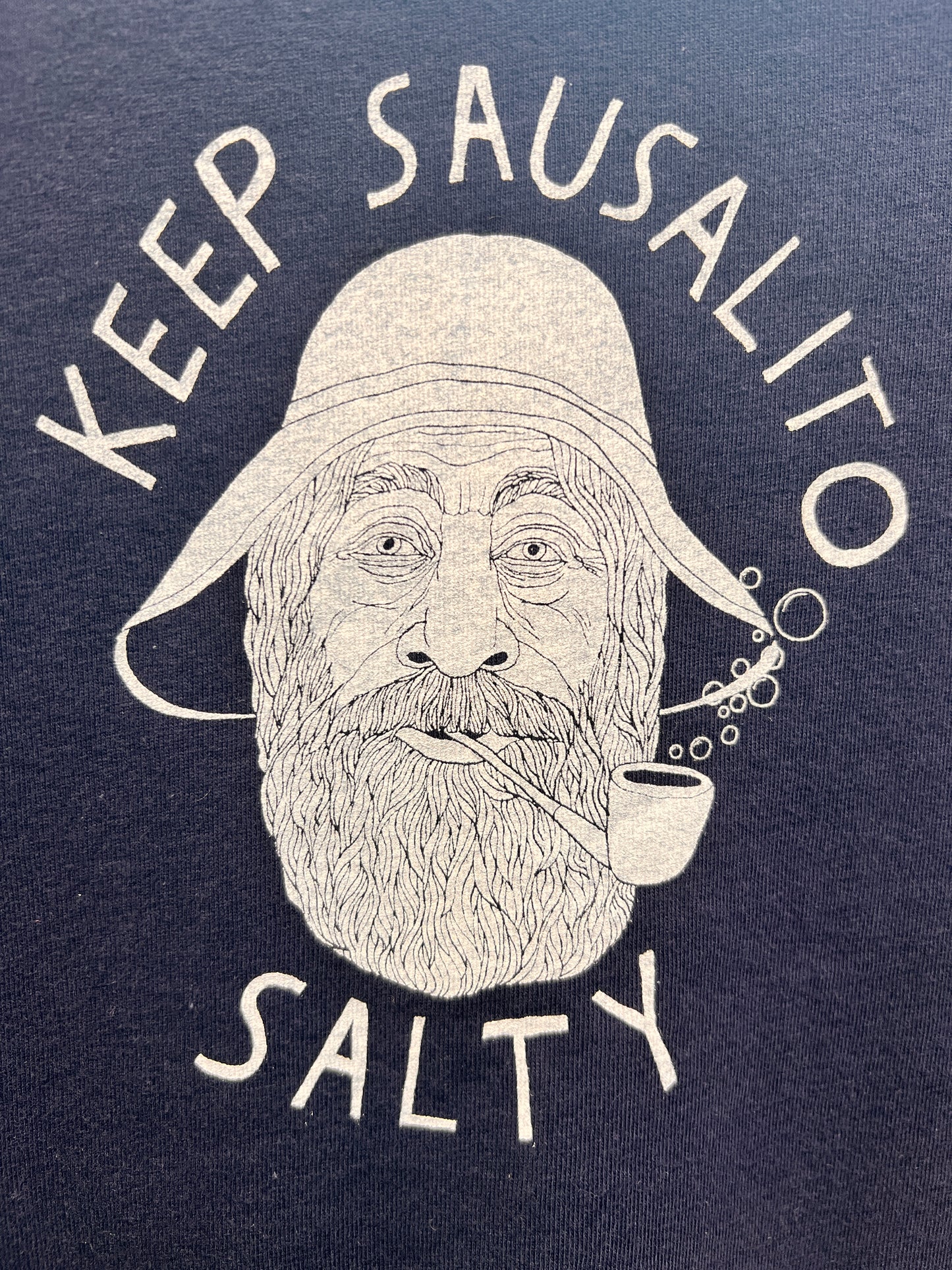Toddler-sized navy blue t-shirt featuring a white graphic of a bearded sailor in a rain hat with 'Keep Sausalito Salty' text and a bubble pipe blowing bubbles.