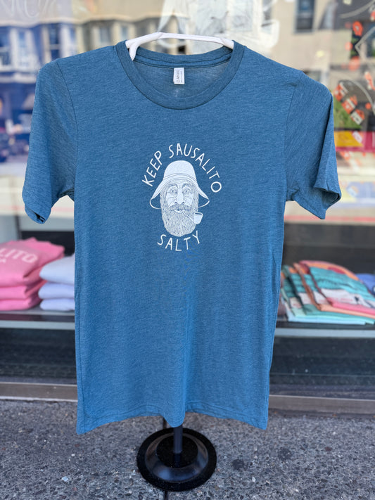Front view of a steel blue, heathered crewneck t-shirt displayed on a black tabletop clothing mannequin stand in front of a shop window. The shirt features a white circular graphic screen-printed on the center chest, showcasing a detailed illustration of a classic, bearded coastal fisherman wearing a traditional weather hat and smoking a pipe. The text "KEEP SAUSALITO" arches cleanly over the top of the portrait, and the word "SALTY" curves directly underneath.
