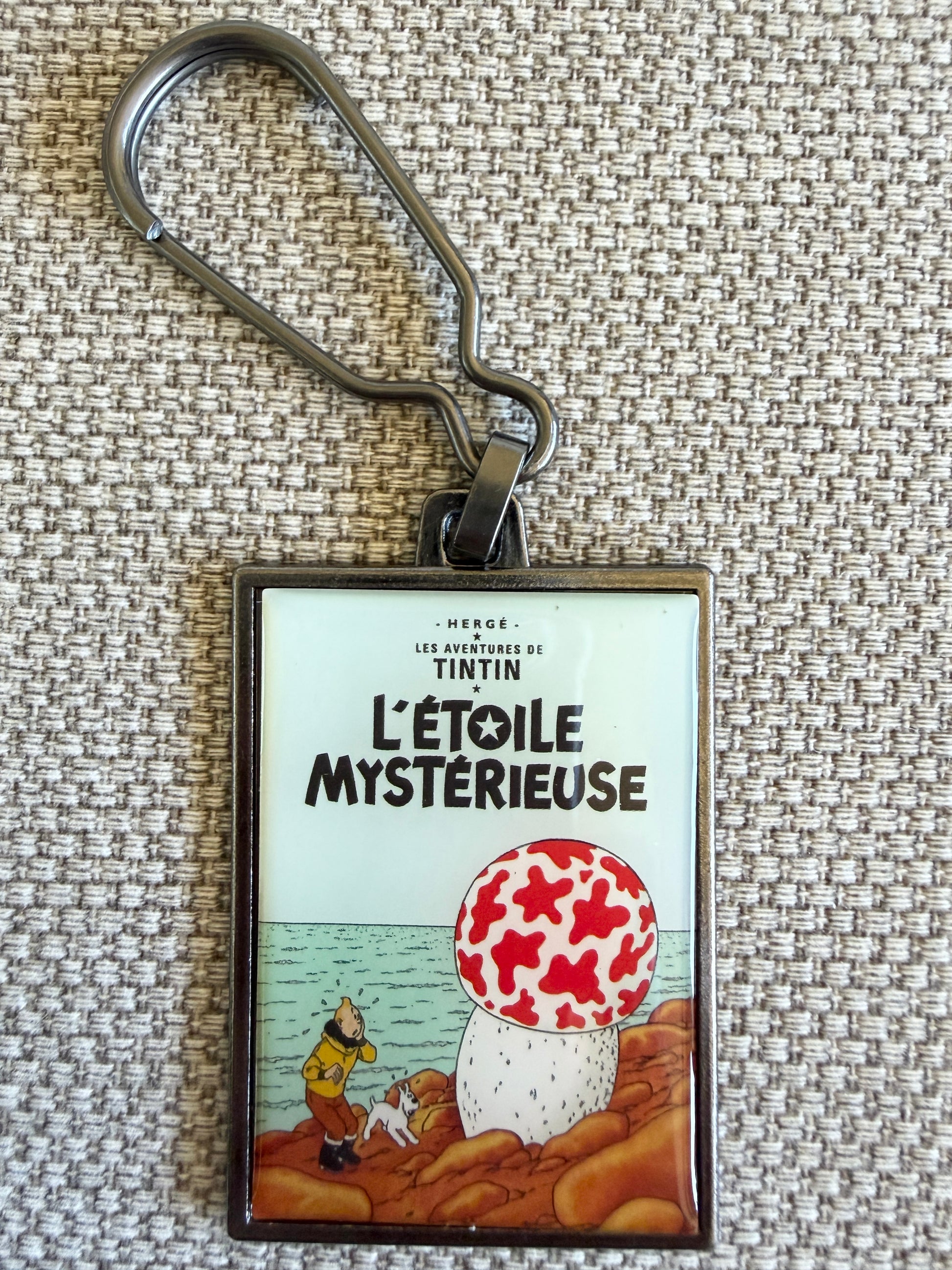 A close-up front view of an officially authorized, rectangular metal Tintin keyring resting on a textured fabric surface. The crisp, resin-coated image reproduces the cover of "The Shooting Star," showing a wide-eyed Tintin and Snowy standing on an uneven, rocky brown landscape next to a massive, surreal white mushroom covered in large red spots under a dark blue sky.