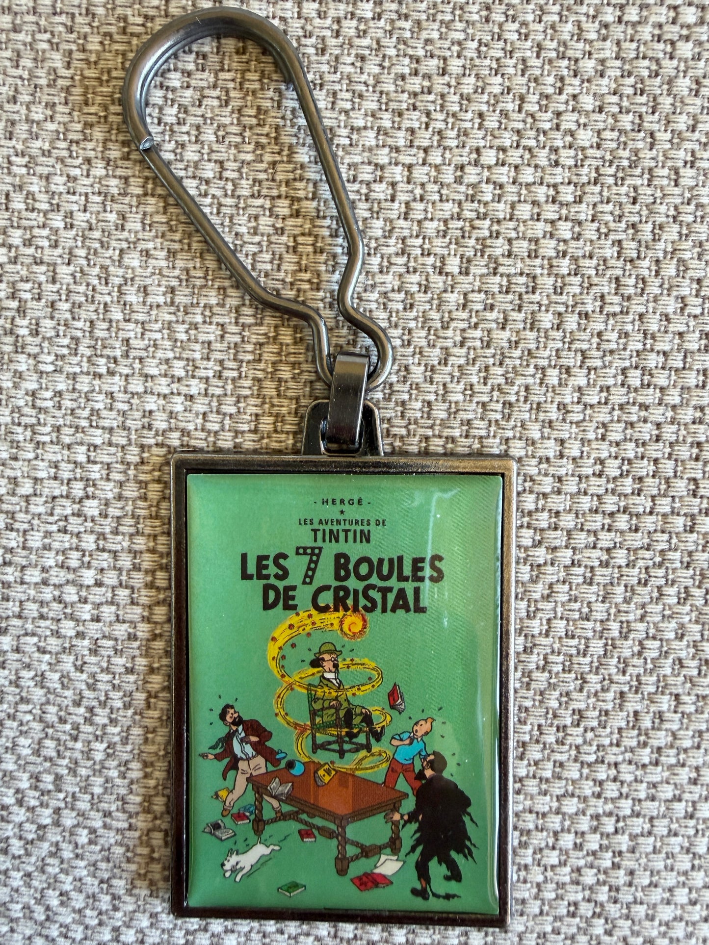 A close-up front view of an officially authorized, rectangular metal Tintin keyring resting on a textured, light-colored fabric background. The glossy, resin-coated image showcases the dramatic cover of "The Seven Crystal Balls," featuring a startled Tintin and Snowy peering out from the dark as a bright yellow lightning bolt strikes across a deep teal sky behind Professor Tarragon's mansion.