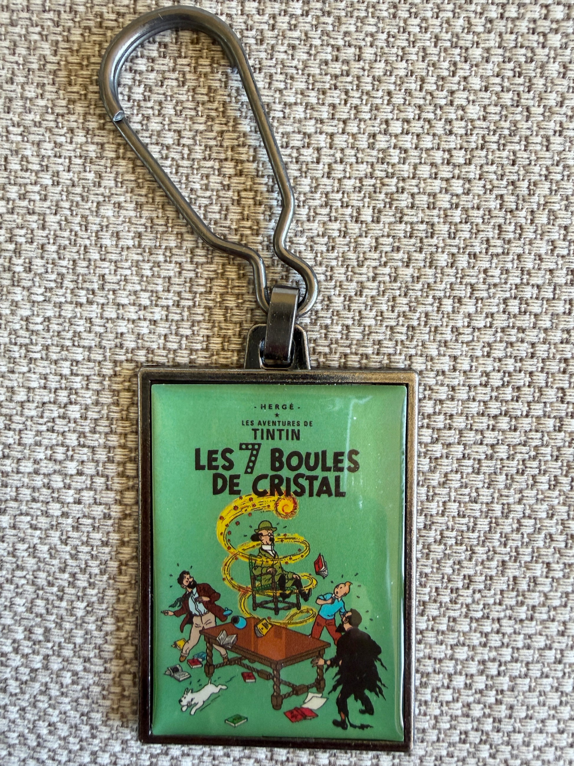 A close-up front view of an officially authorized, rectangular metal Tintin keyring resting on a textured, light-colored fabric background. The glossy, resin-coated image showcases the dramatic cover of "The Seven Crystal Balls," featuring a startled Tintin and Snowy peering out from the dark as a bright yellow lightning bolt strikes across a deep teal sky behind Professor Tarragon's mansion.