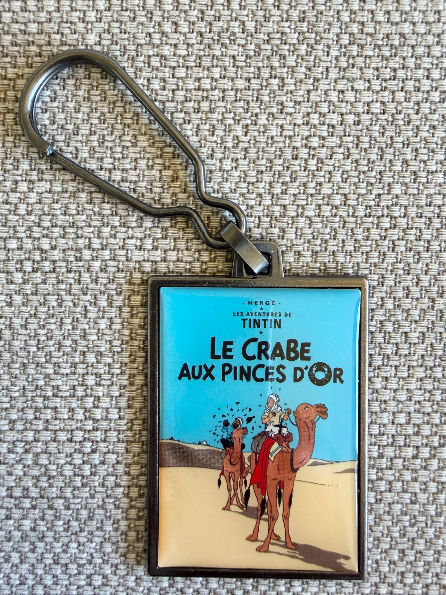 A rectangular keychain displaying the French book cover art for Hergé's Tintin adventure "Le Crabe aux pinces d'or." The illustration shows Tintin and Captain Haddock trekking across vast, rolling desert dunes under a warm golden sky, accompanied by a caravan of camels.