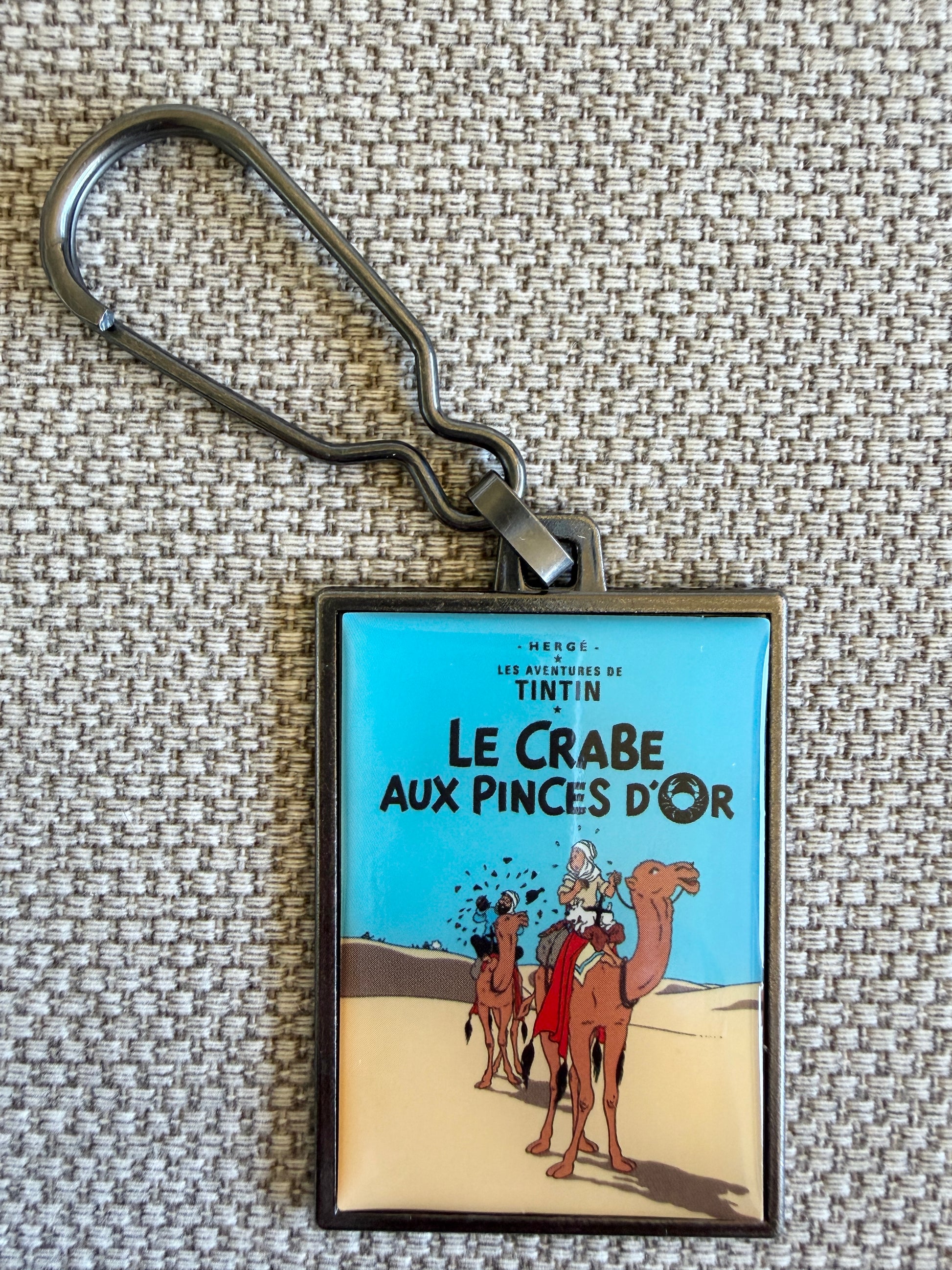 A rectangular keychain displaying the French book cover art for Hergé's Tintin adventure "Le Crabe aux pinces d'or." The illustration shows Tintin and Captain Haddock trekking across vast, rolling desert dunes under a warm golden sky, accompanied by a caravan of camels.
