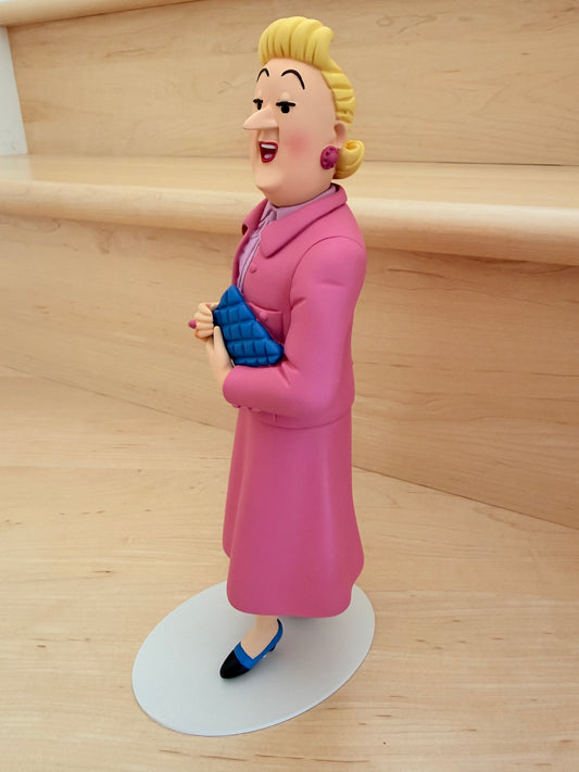 Side profile view of the premium hand-painted resin figurine of Madame Bianca Castafiore from the Tintin Icons collection. The 26cm tall statue features the character standing elegantly on a white oval base over a light wood surface, wearing her signature bright, rosy pink winter coat and a soft lavender collared shirt. Her blonde hair is styled in her classic updos, and she is holding a textured, deep blue quilted clutch bag close to her waist.