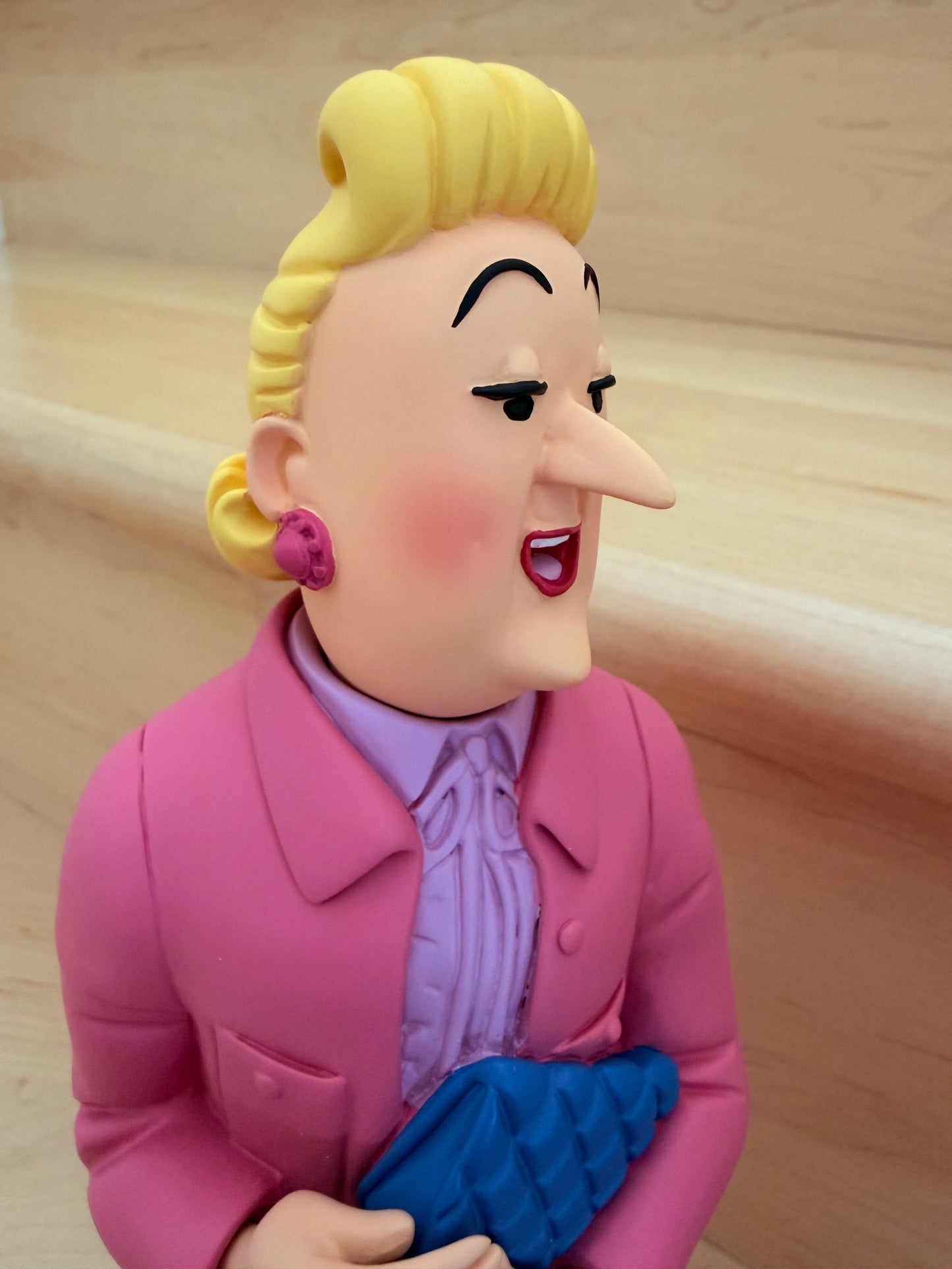 Side-profile close-up view of the premium hand-painted resin figurine of Madame Bianca Castafiore from the Tintin Icons collection. The statue captures her from the torso up, showcasing her prominent nose, styled blonde updos, and a detailed magenta rose earring. She wears a bright, rosy pink winter coat over a lavender high-collared blouse with a ruffled tie, while holding her textured, deep blue quilted clutch bag near her waist against a light wooden background.
