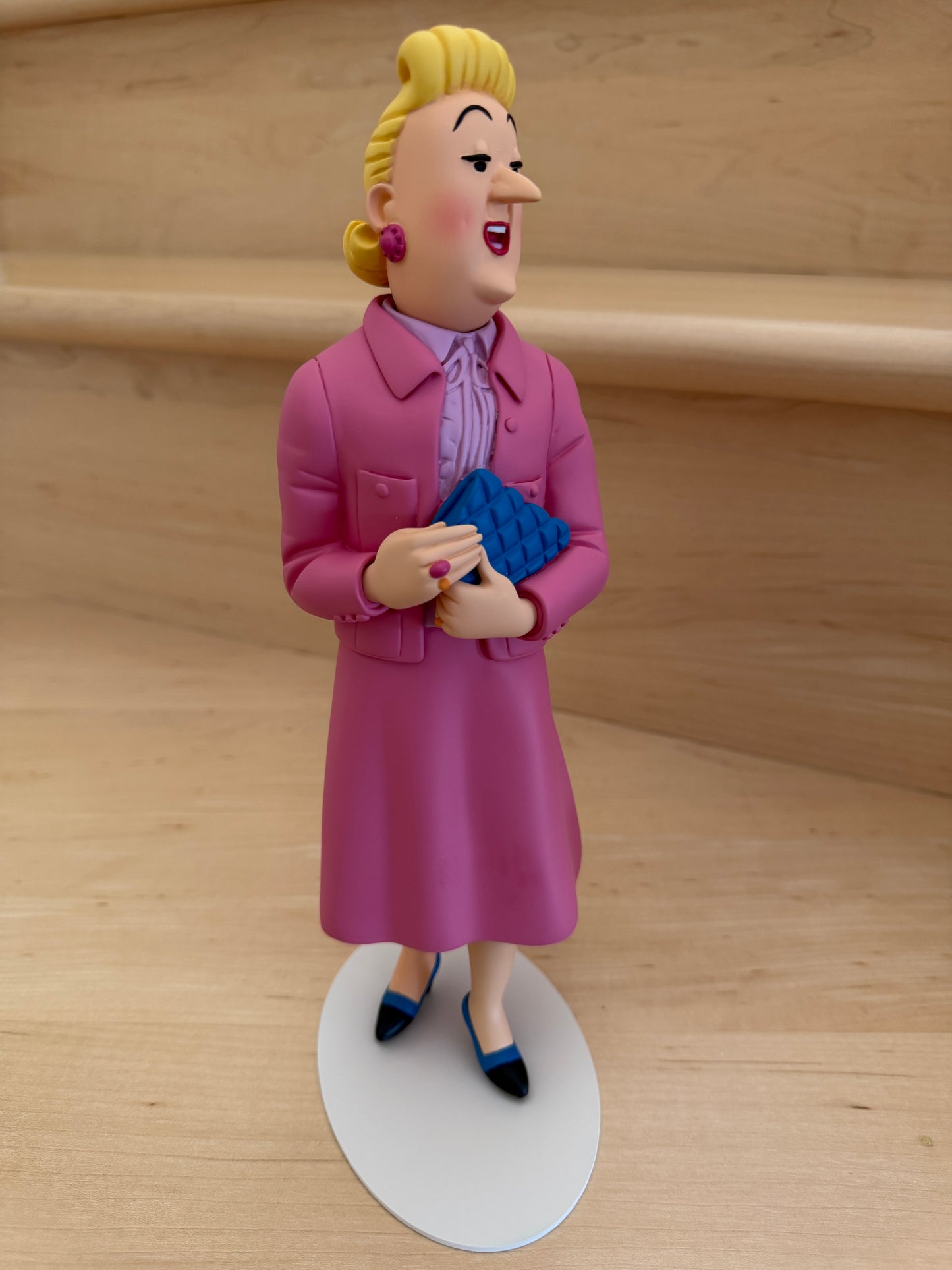 Three-quarter front view of the premium hand-painted resin figurine of Madame Bianca Castafiore from the Tintin Icons collection. The 26cm tall statue depicts the character standing elegantly on a flat, white oval base, wearing her signature bright, rosy pink winter coat over a light lavender collared blouse. She has a vibrant smile, classic blonde updos with matching pink earrings, and is holding a textured, deep blue quilted clutch bag with both hands near her waist.