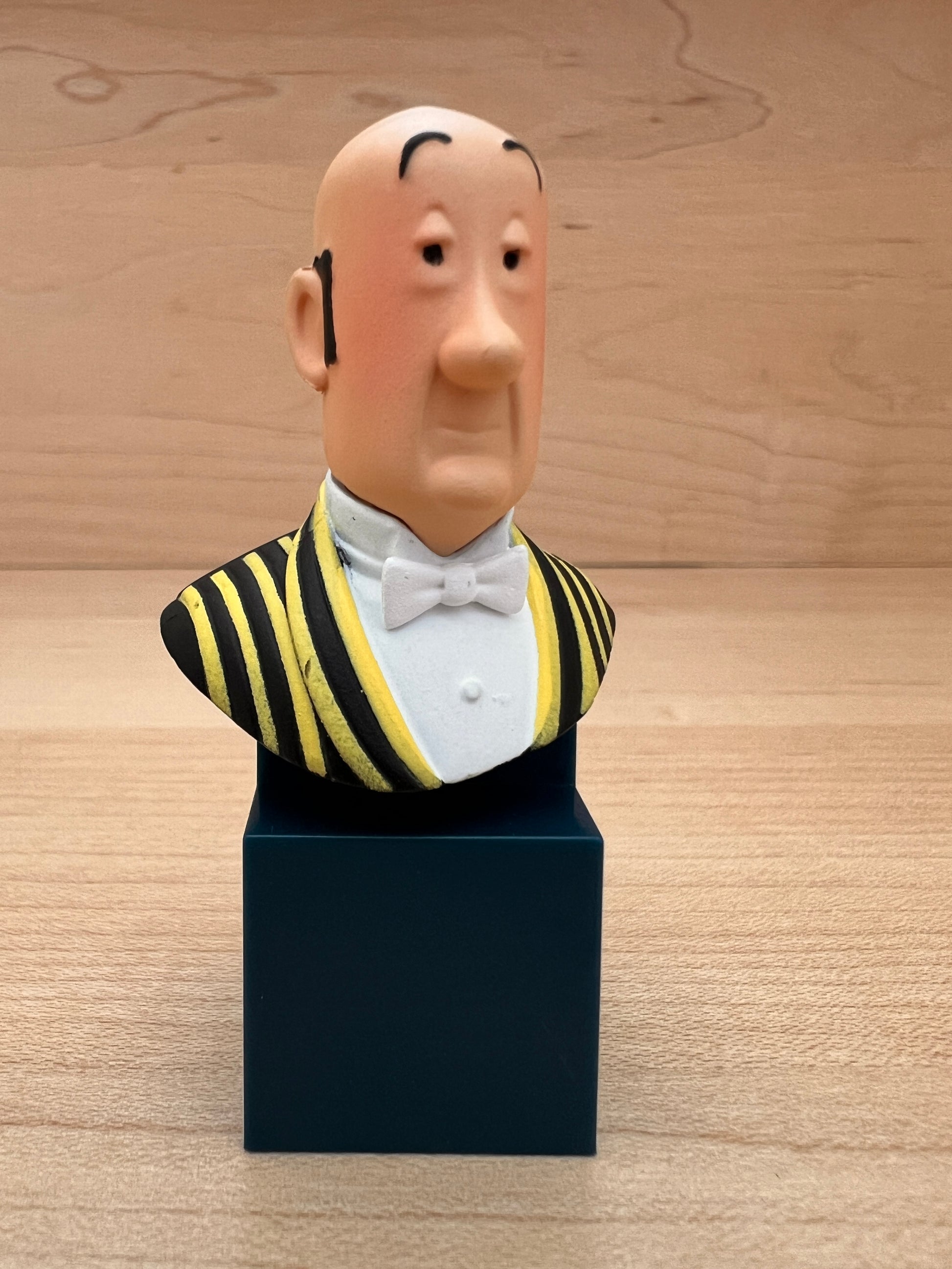 A mini PVC collectible bust figure of Nestor, Captain Haddock's loyal butler from Tintin, wearing a black and yellow striped waistcoat with a white bow tie, mounted on a dark blue square pedestal.